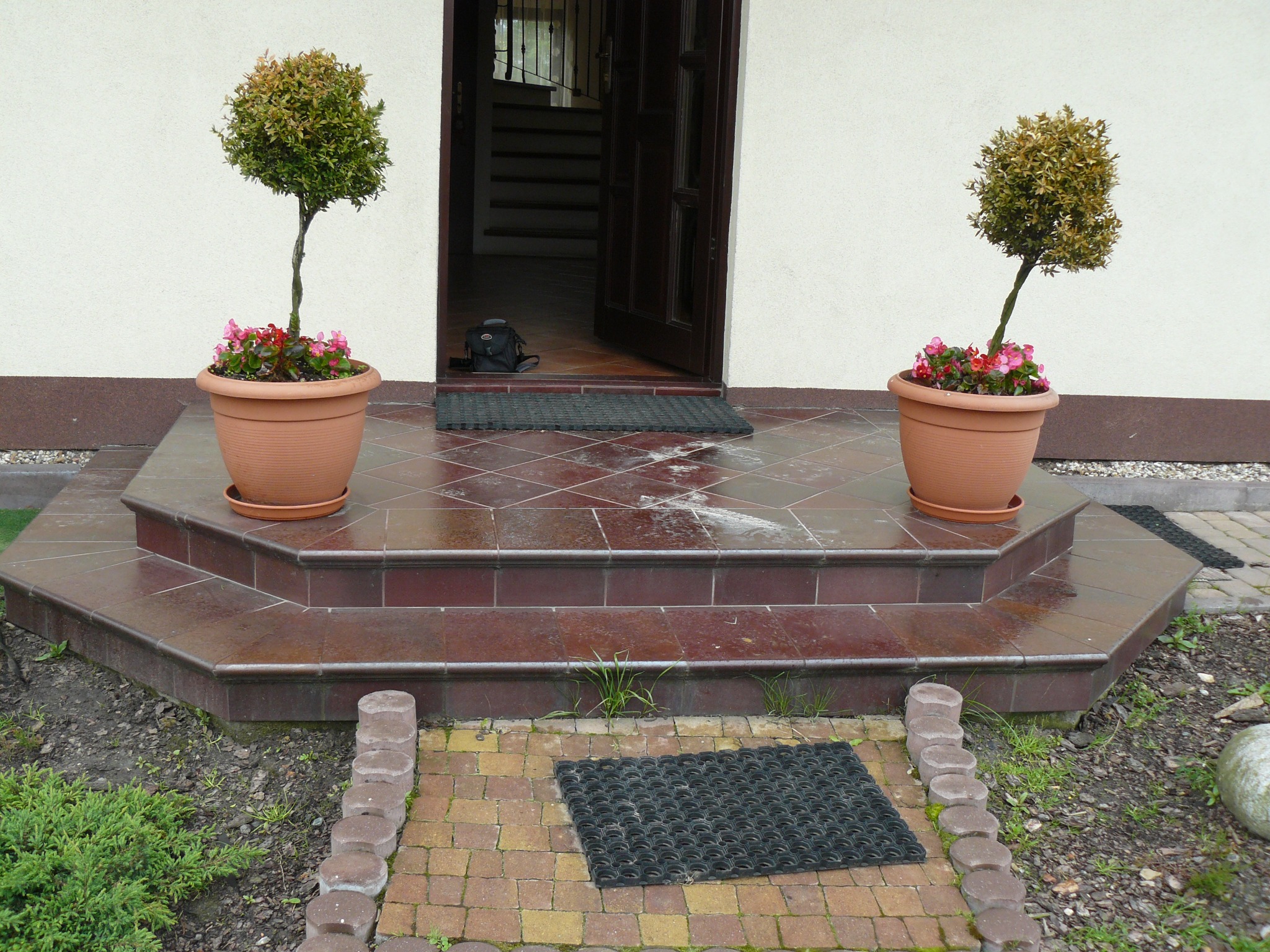 Wykończone płytkami schody wejściowe z dwoma donicami z drzewkami po bokach. Widoczne brązowe płytki i ceglana ścieżka prowadząca do drzwi wejściowych.