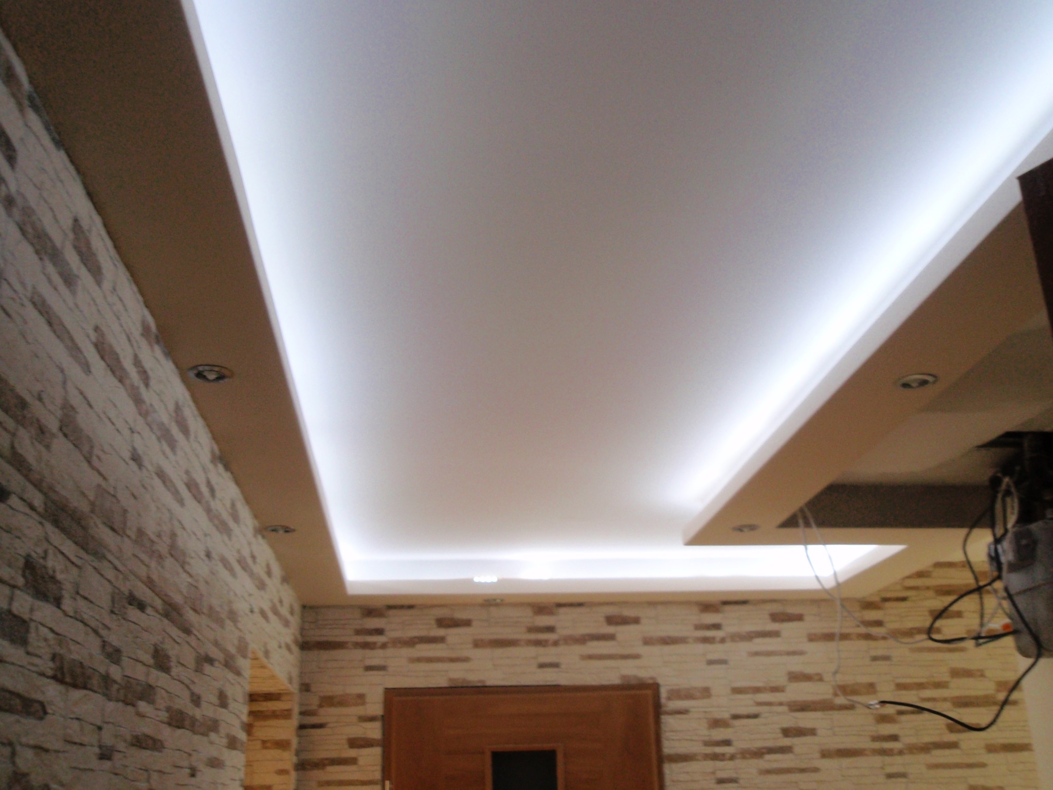 Podwieszany sufit z oświetleniem LED i dekoracyjna ściana z imitacji kamienia. Widoczne elementy instalacji elektrycznej. Wykończenie wnętrza w ciepłych barwach.
