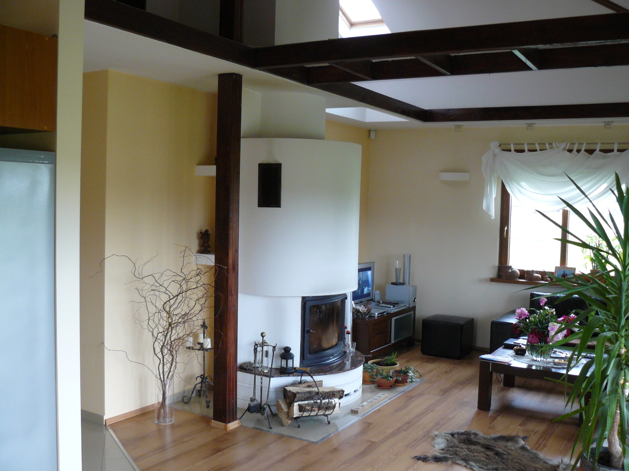 Jasne wnętrze z kominkiem o zaokrąglonym kształcie, drewnianą podłogą i elementami dekoracyjnymi. Widoczne okno z firanką i rośliny doniczkowe.