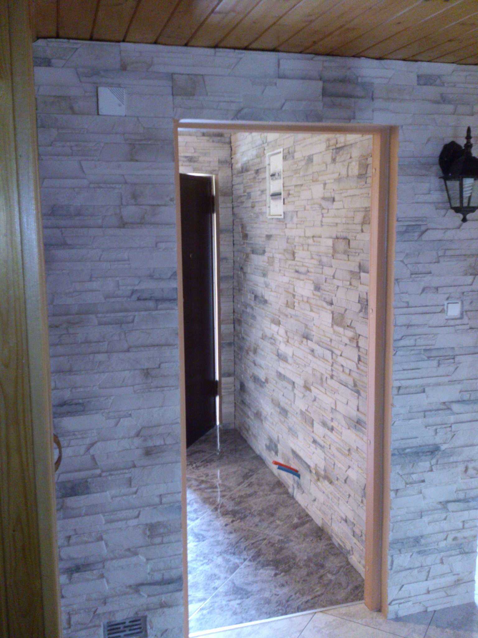 Wykończenie korytarza z dekoracyjną okładziną kamienną na ścianach i brązową podłogą z płytek. Widoczna drewniana boazeria sufitowa i lampa ścienna.