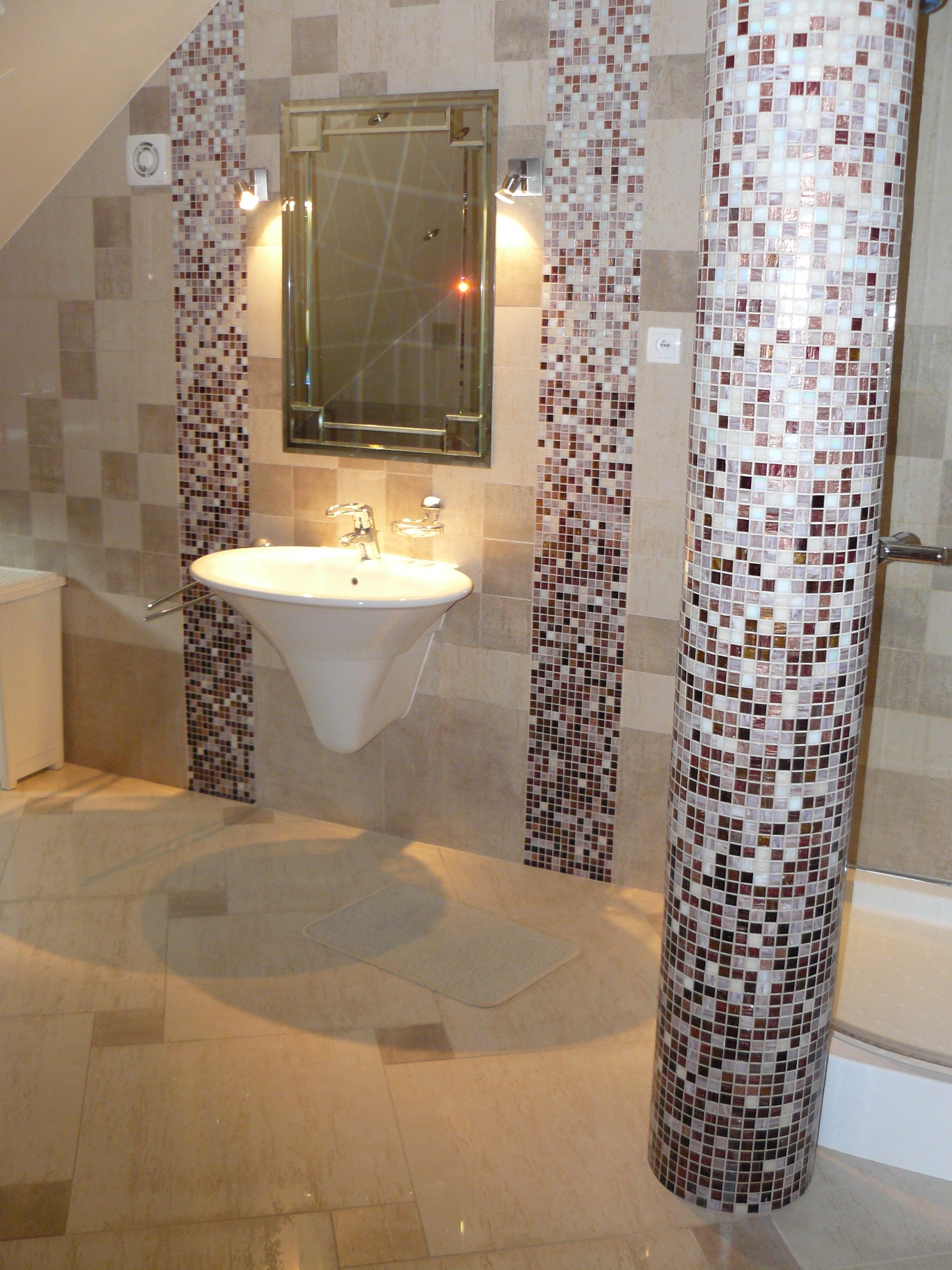 Wykończona łazienka z mozaiką w odcieniach brązu i beżu. Umywalka wisząca, lustro w ramie i kolumna obłożona mozaiką. Jasne płytki podłogowe.