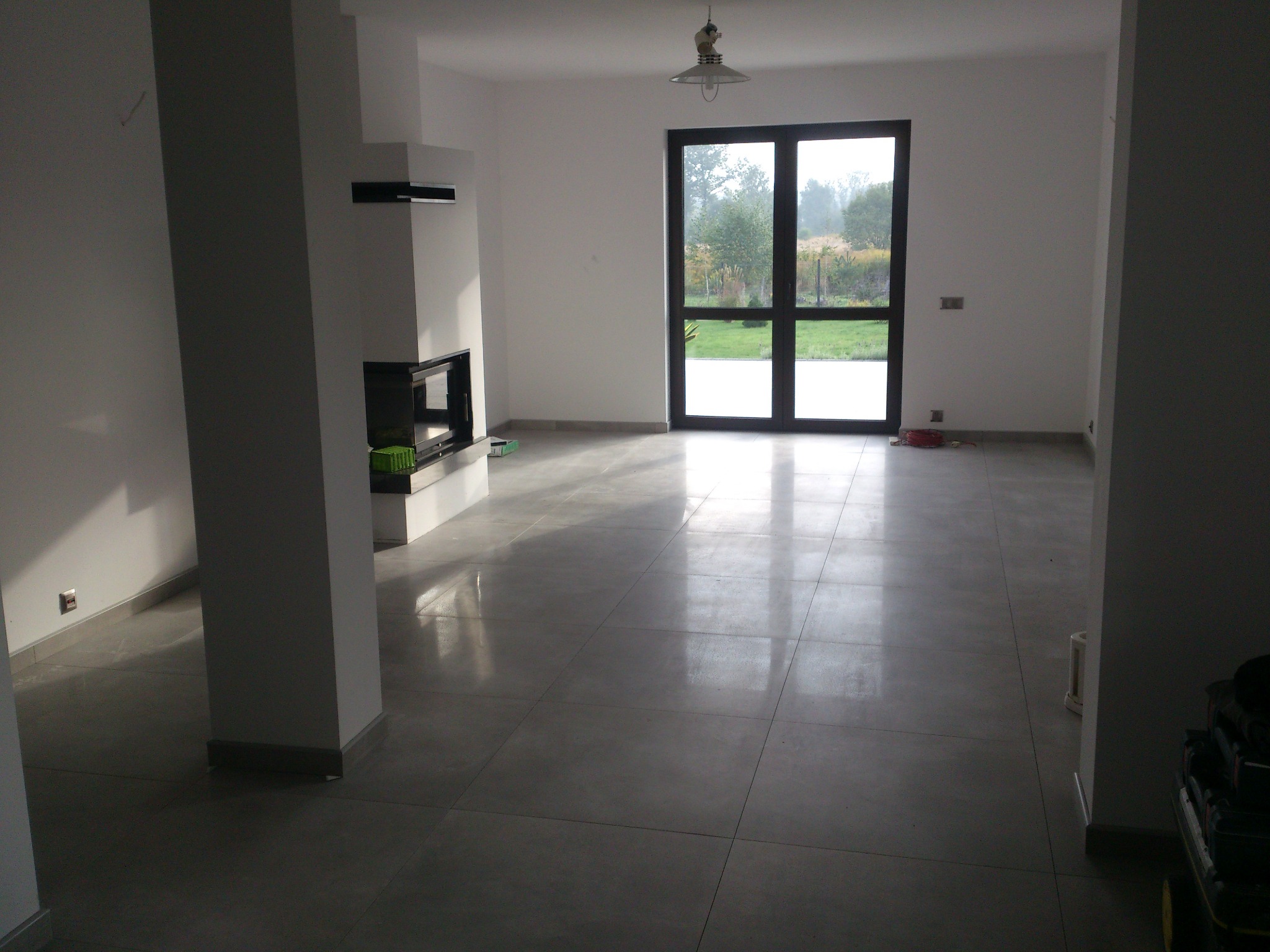 Jasne, przestronne wnętrze po remoncie w Sosnowcu. Szare płytki podłogowe, białe ściany, nowoczesny kominek i duże okno z widokiem na zieleń.