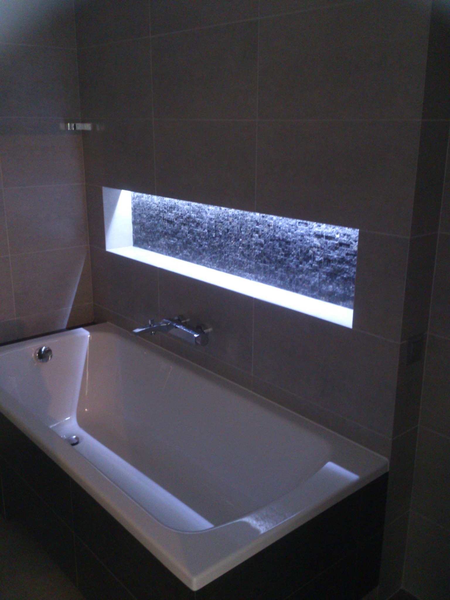Nowoczesna łazienka z wanną obudowaną ciemnymi płytkami i wnęką z dekoracyjną mozaiką podświetloną ledami, wykończona w szarościach.