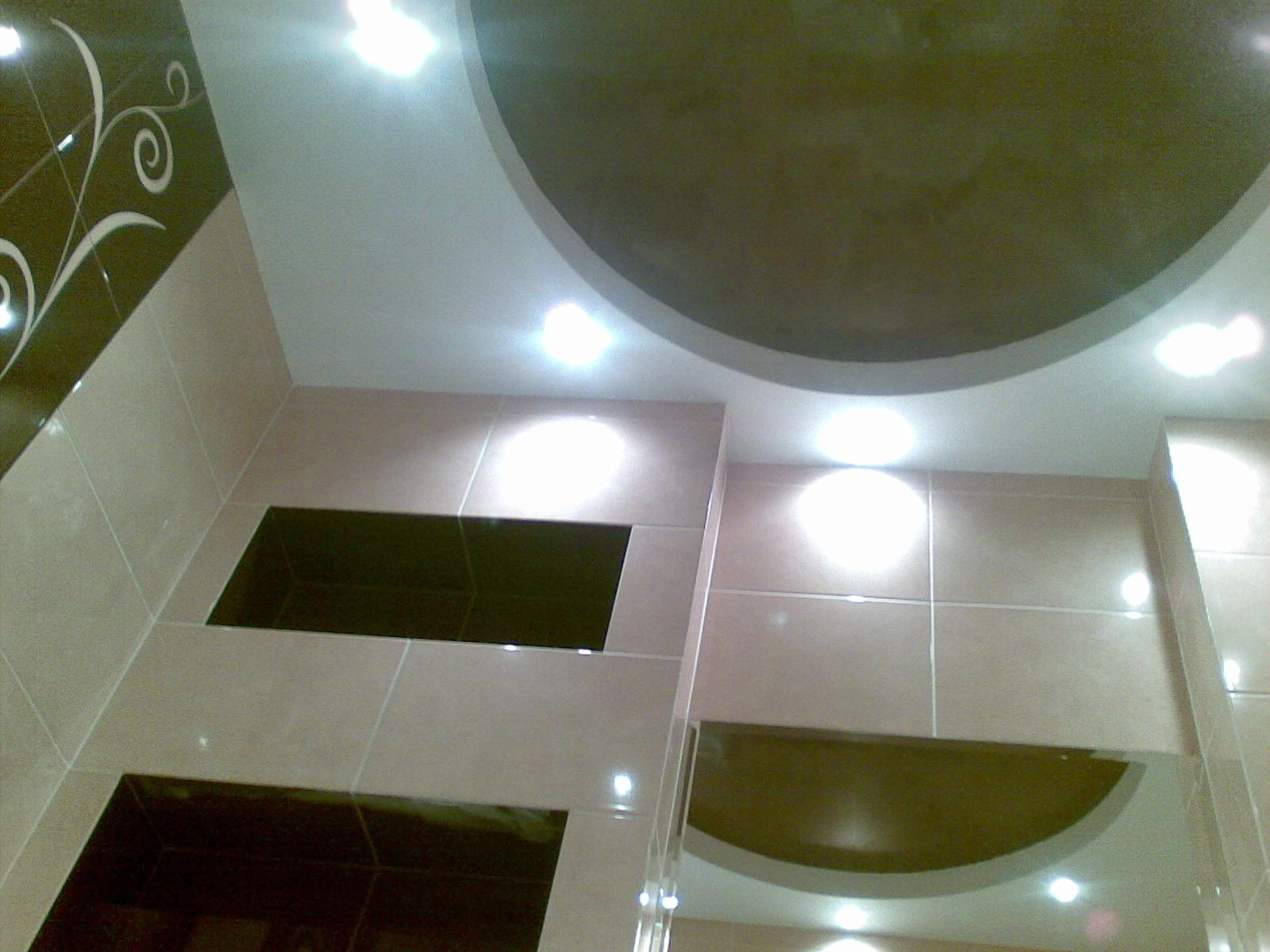 Wykończona łazienka z beżowymi płytkami, dekoracyjnym wzorem na ścianie i podwieszanym sufitem z okrągłym elementem. Widoczne oświetlenie punktowe.