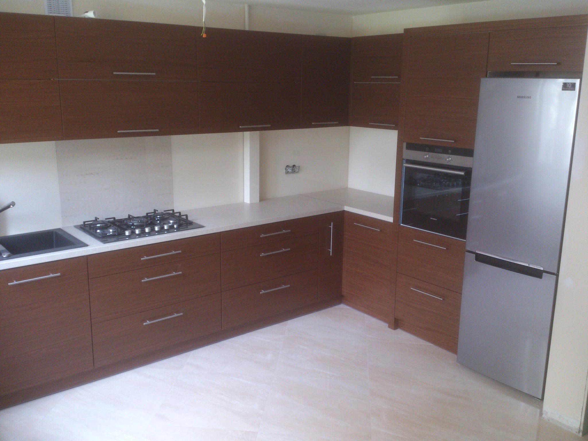 Nowoczesna kuchnia z brązowymi szafkami, jasnym blatem i płytkami podłogowymi. Widoczna płyta gazowa, zlewozmywak i lodówka Samsung. Minimalistyczny design.