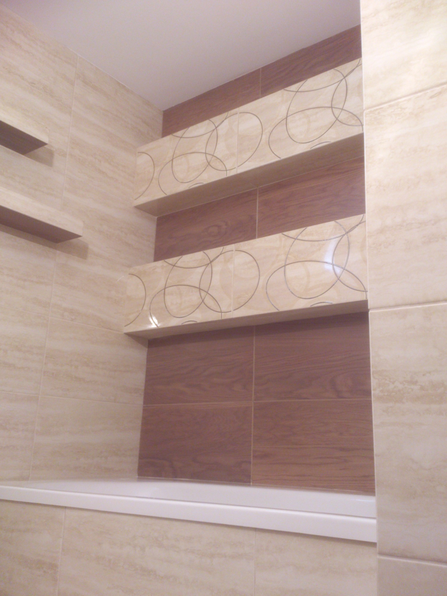 Wykończona łazienka z beżowymi i brązowymi płytkami, nad wanną półki z dekoracyjnym wzorem. Widok na fragment ściany i wanny, bez armatury.