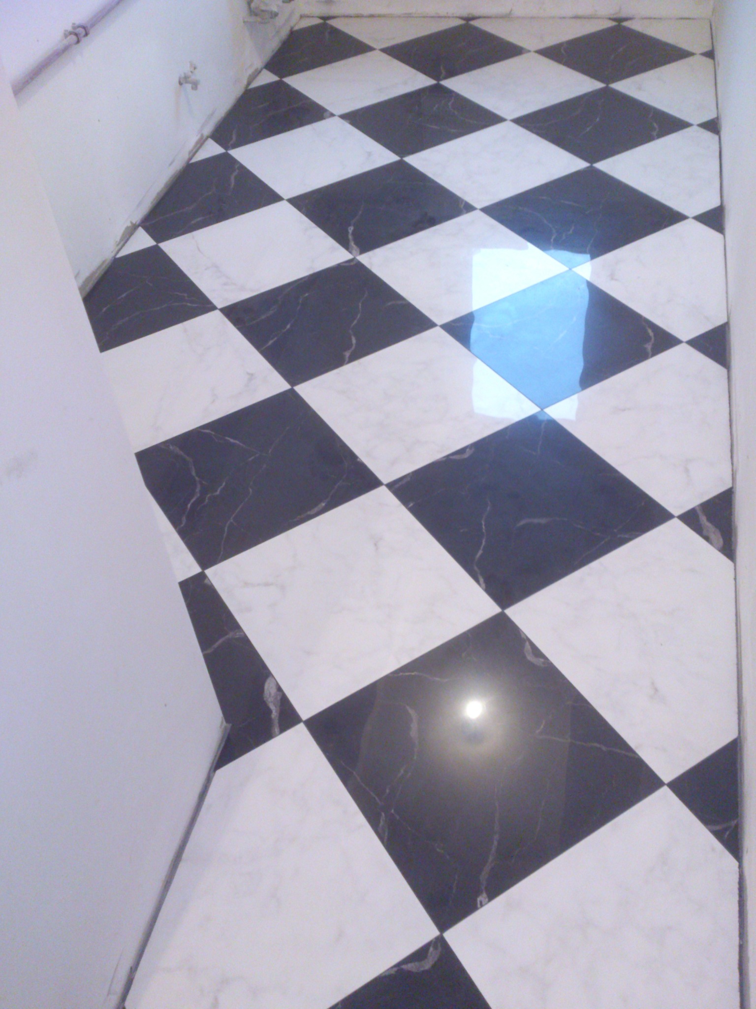 Ukończona podłoga w szachownicę z czarno-białych płytek marmurowych, ułożonych pod kątem w małej łazience. Widoczne odbicie światła na powierzchni płytek.