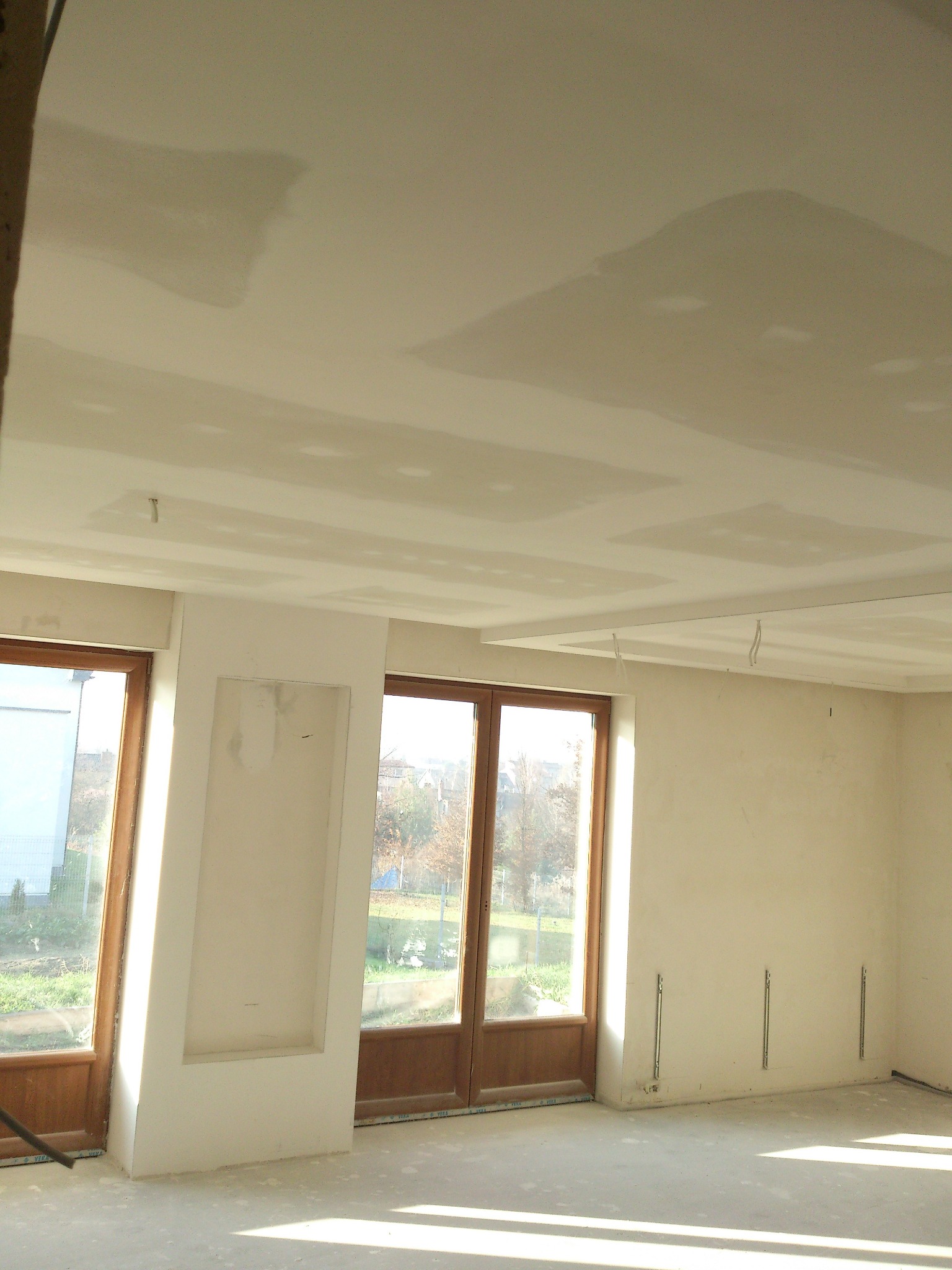 Wnętrze w trakcie szpachlowania: gładkie ściany i sufit, drewniane okna z widokiem na zewnątrz, przygotowane do dalszych prac wykończeniowych. Jasne światło dzienne.