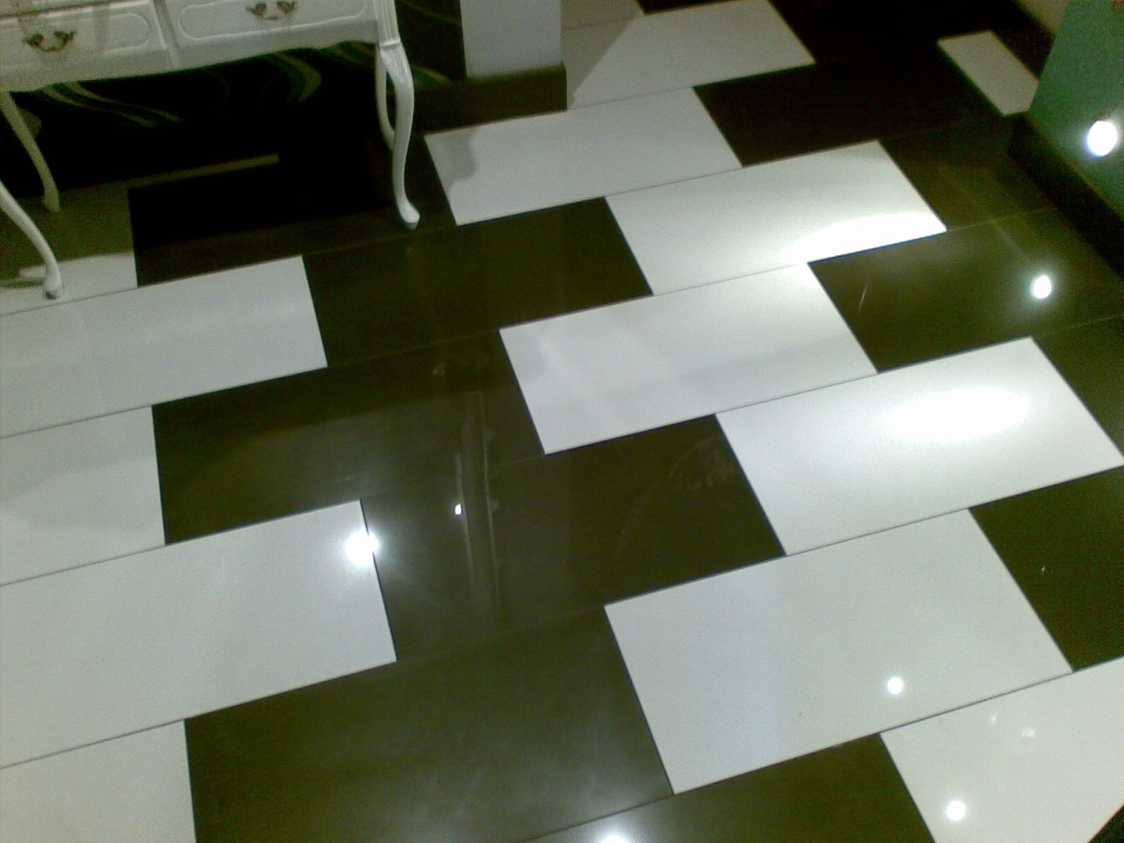 Ukończona podłoga z błyszczących, prostokątnych płytek w szachownicę (biel i ciemny brąz), widoczny fragment białej komody na nóżkach.