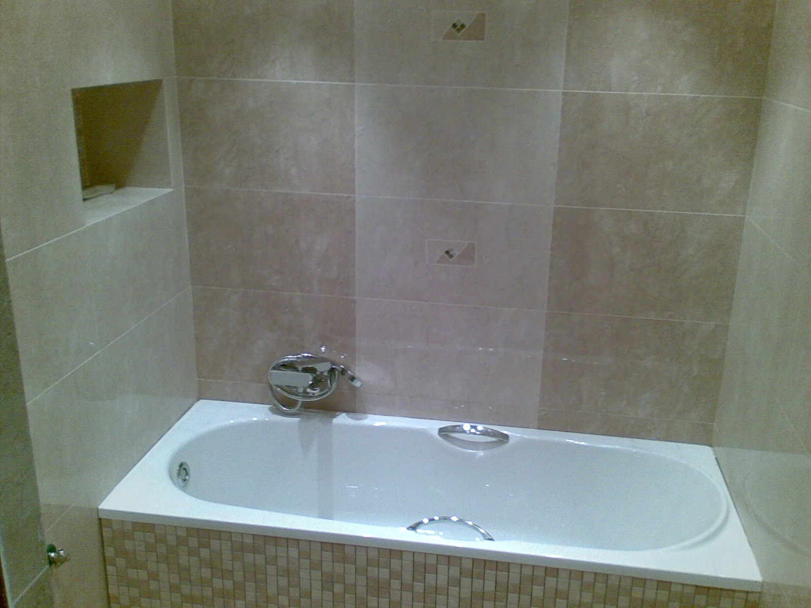 Wykończona łazienka z wanną obłożoną mozaiką i beżowymi płytkami z dekorami. W ścianie wnęka. Bateria wannowa chromowana. Widok na fragment ściany i wannę.