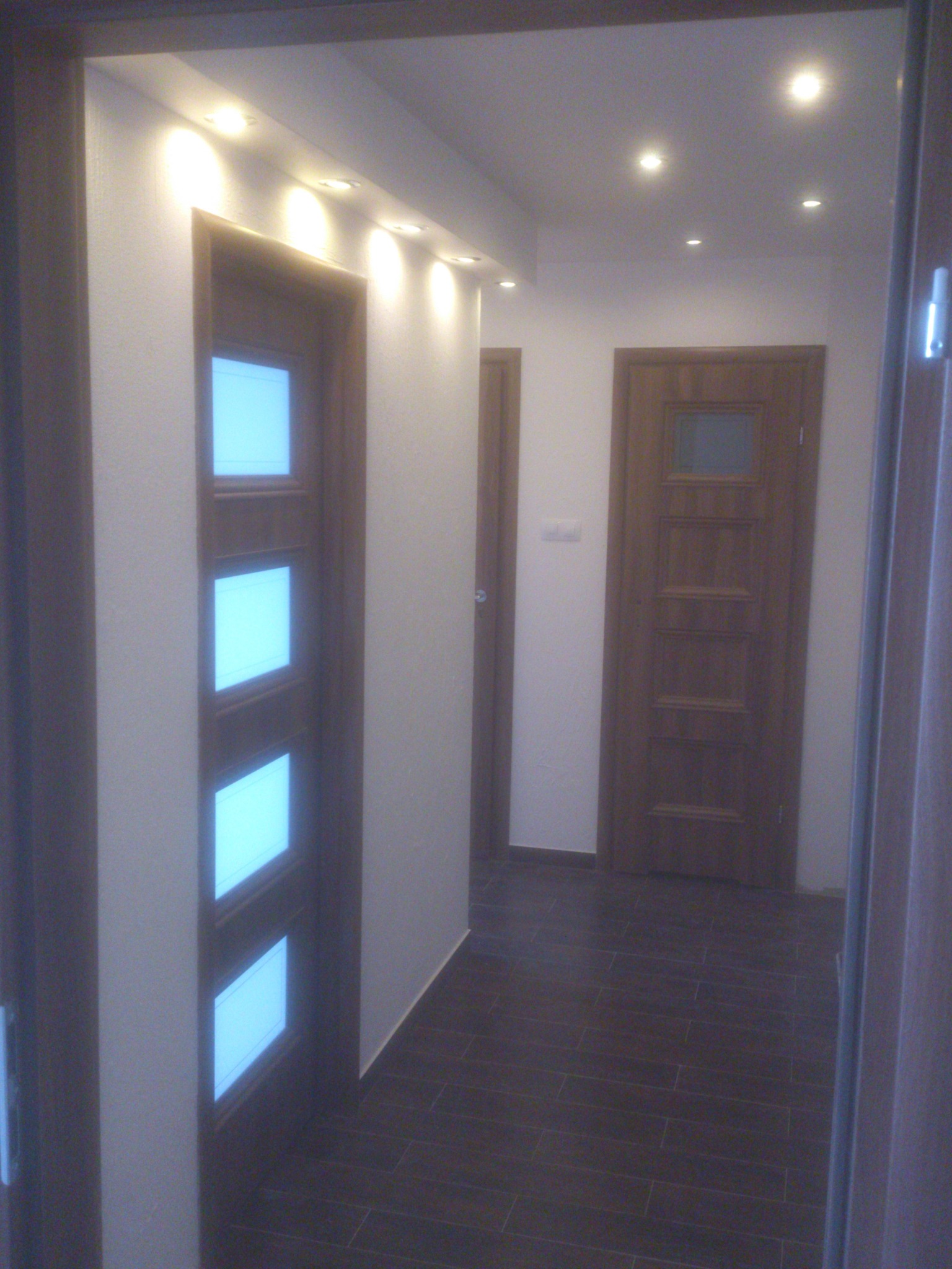 Korytarz z dwiema drewnianymi drzwiami, jedna z czterema przeszkleniami, oświetlenie punktowe sufitowe, ciemna podłoga z płytek. Widok z perspektywy.