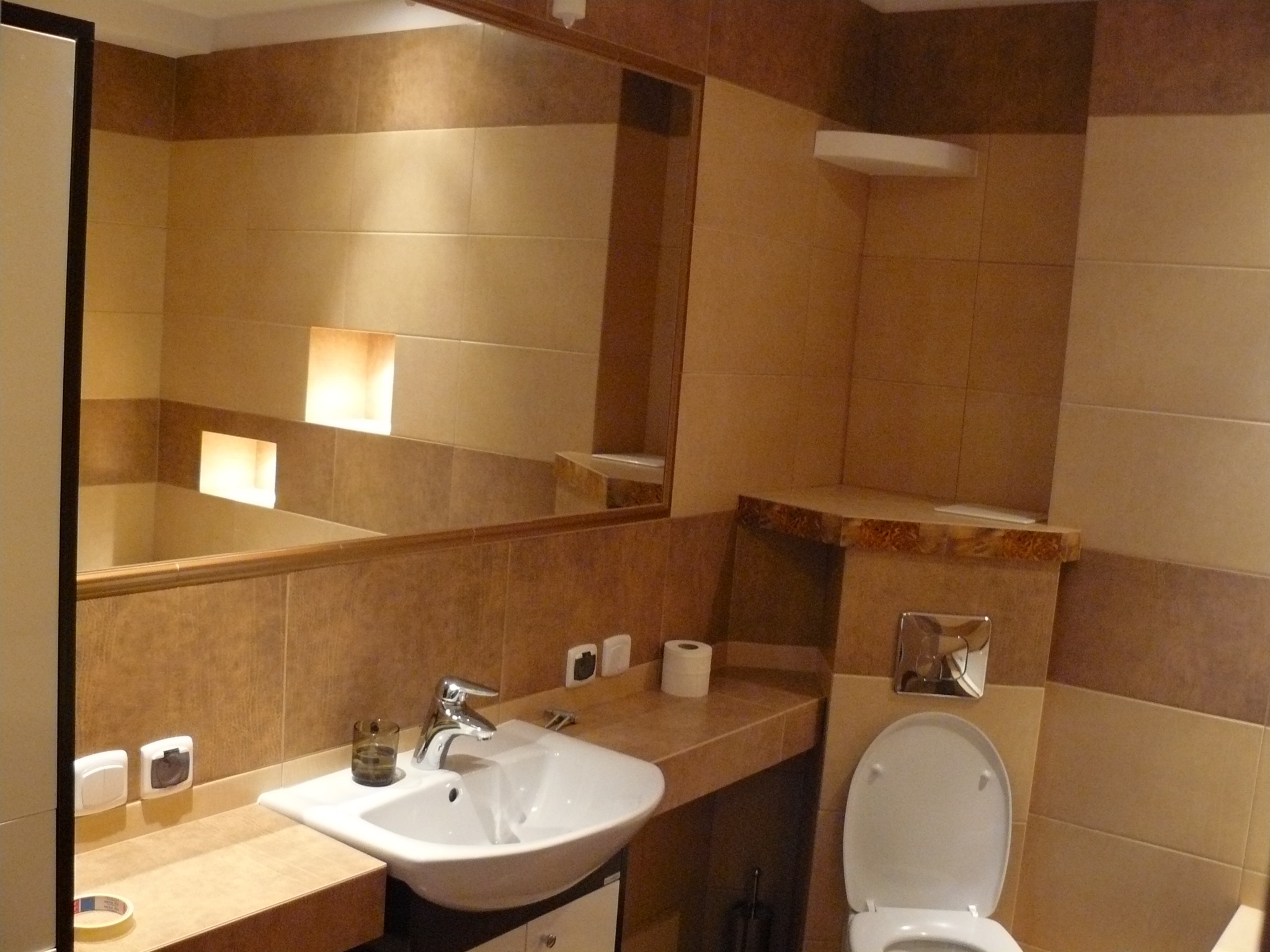 Wykończona łazienka w ciepłych brązach z umywalką, toaletą i dużym lustrem. Widoczne płytki w dwóch odcieniach brązu, oświetlenie wnękowe i akcesoria łazienkowe.