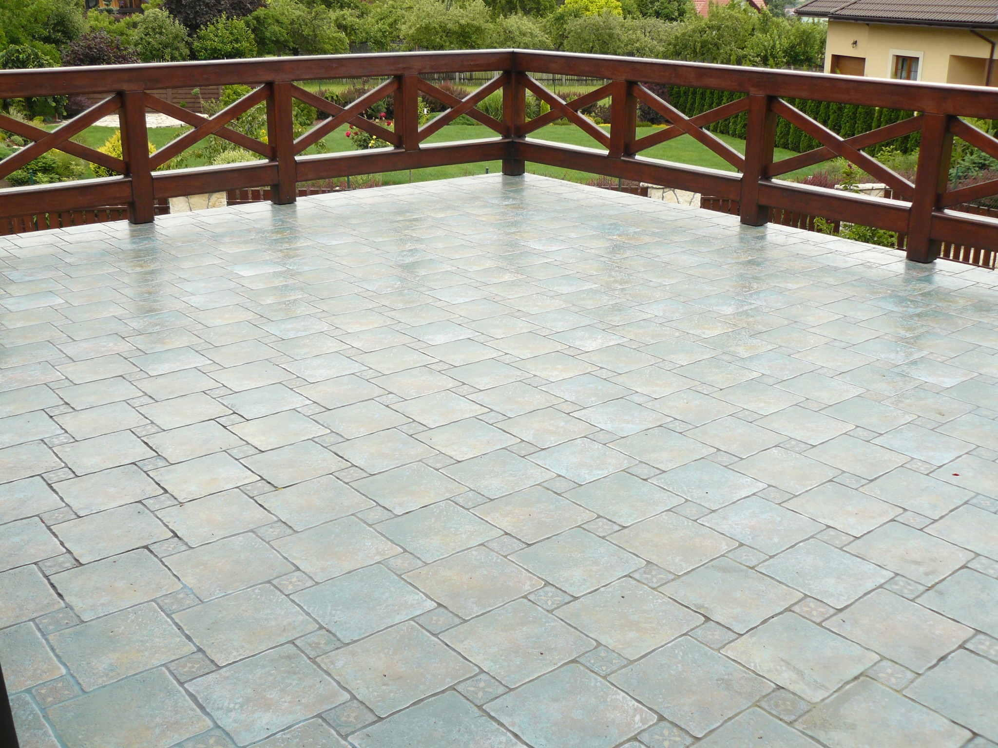 Wykończony balkon z drewnianą balustradą i nową, jasną, kwadratową terakotą ułożoną w karo. Widok na zieleń ogrodu w tle.