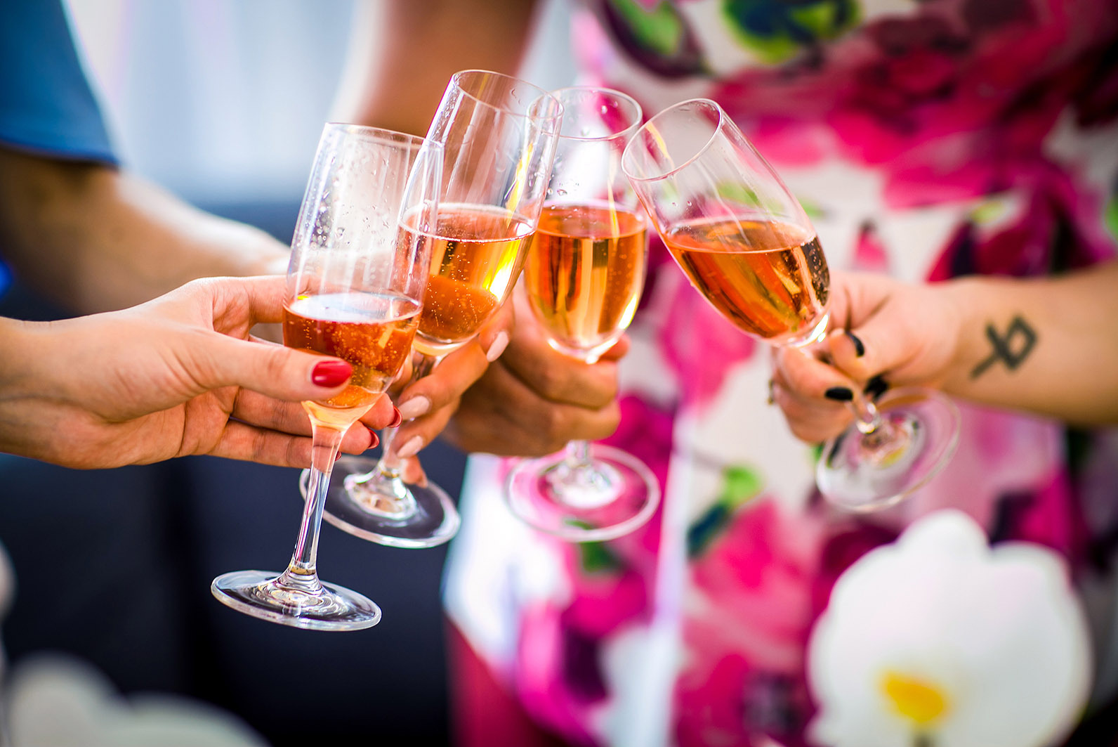Cztery osoby wznoszą toast musującym napojem w smukłych kieliszkach. W tle kwiatowy wzór sukienki i tatuaż na nadgarstku. Jasne, naturalne światło.