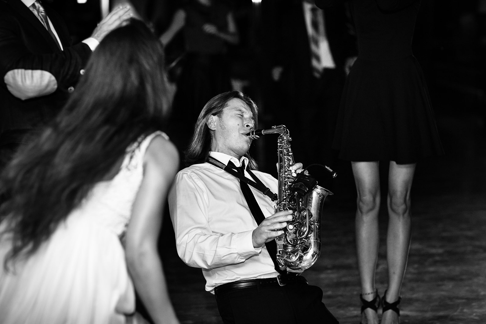 Saksofonista w akcji, czarno-białe zdjęcie z imprezy. Muzyk w białej koszuli i krawacie gra na saksofonie, w tle rozmazane sylwetki tańczących osób.