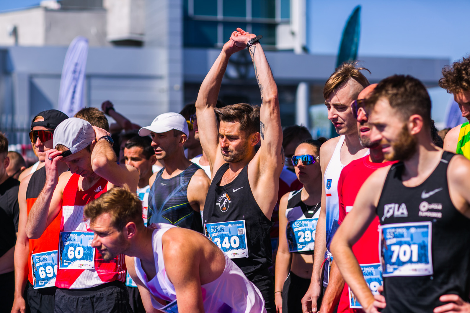 Uczestnicy biegu ulicznego, w sportowych strojach, ustawieni w rzędzie przed startem. Widoczny mężczyzna z uniesionymi rękami i numerem startowym 340.
