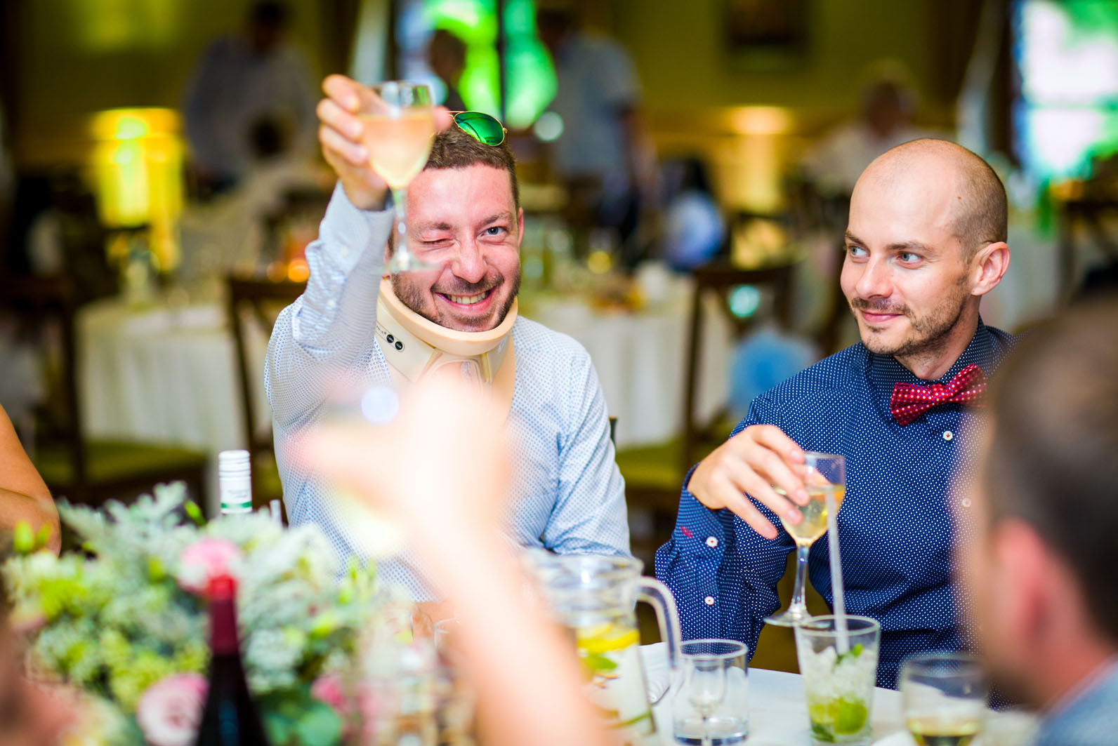 Radosny toast na przyjęciu: mężczyzna w kołnierzu ortopedycznym wznosi kieliszek, drugi z uśmiechem trzyma drinka. Dekoracje stołu i goście w tle.