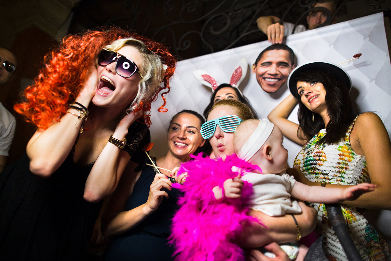 Rozbawiona grupa osób pozuje z rekwizytami w fotobudce podczas imprezy. W tle biała ścianka z wzorem i wystający mężczyzna z maską Obamy.