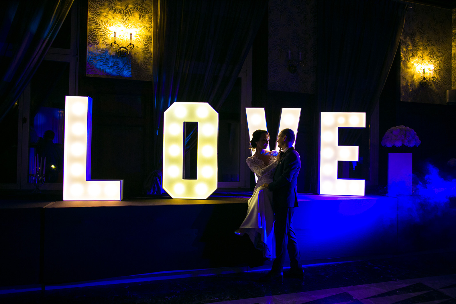Para młoda w objęciach na tle podświetlanych liter 'LOVE' w ciemnym wnętrzu z dekoracjami. Romantyczna atmosfera, subtelne oświetlenie, elegancki strój.