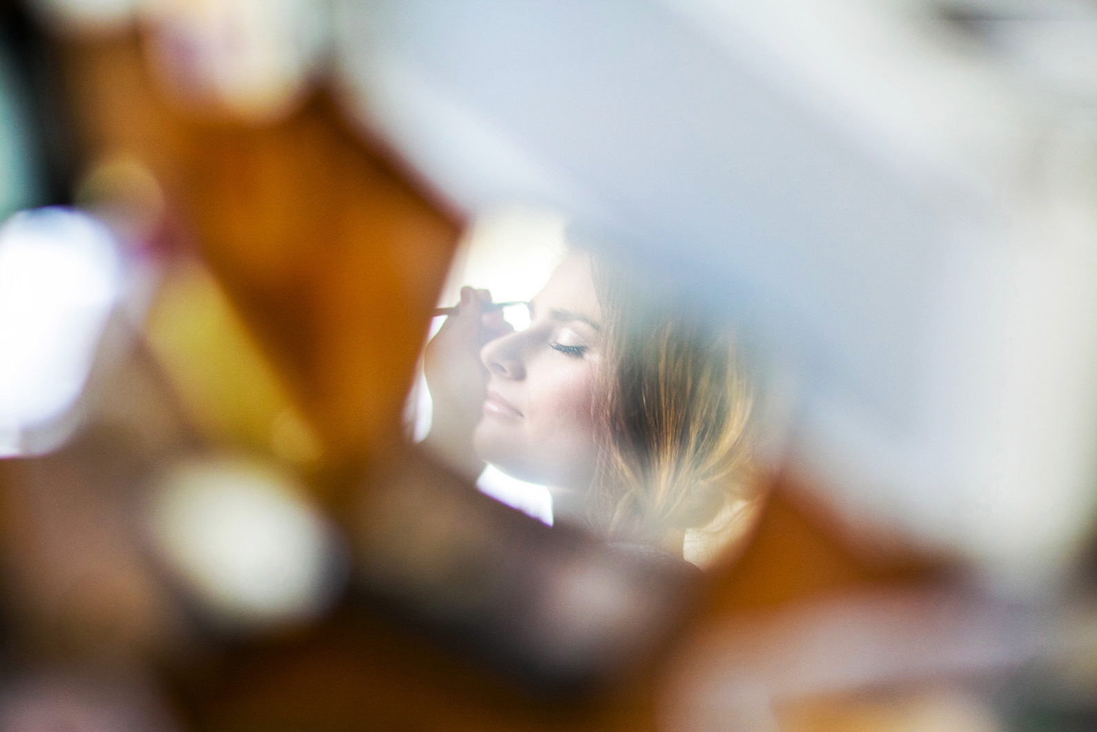 Delikatny portret kobiety z zamkniętymi oczami podczas profesjonalnego makijażu, uchwycony przez pryzmat rozmytego tła, podkreślający subtelność i przygotowania do ważnego wydarzenia.