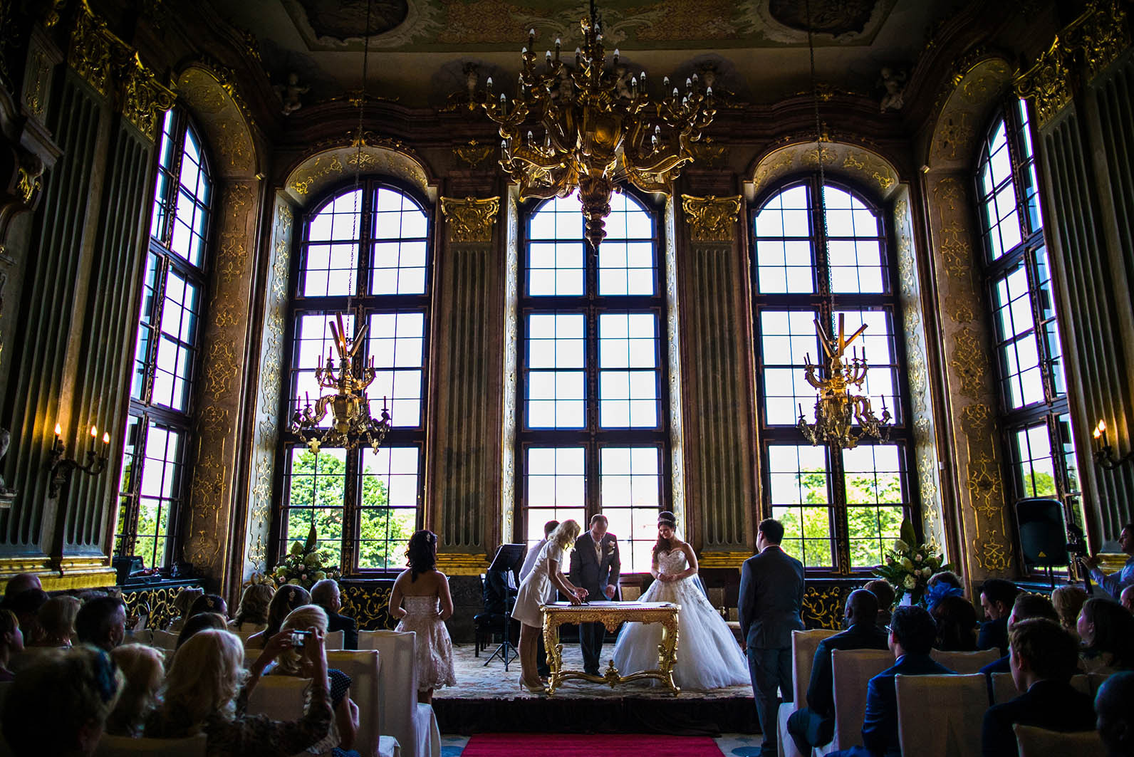 Podpisanie aktu ślubu przez parę młodą w bogato zdobionej sali z wysokimi oknami i żyrandolami. Goście siedzą na krzesłach, obserwując ceremonię.