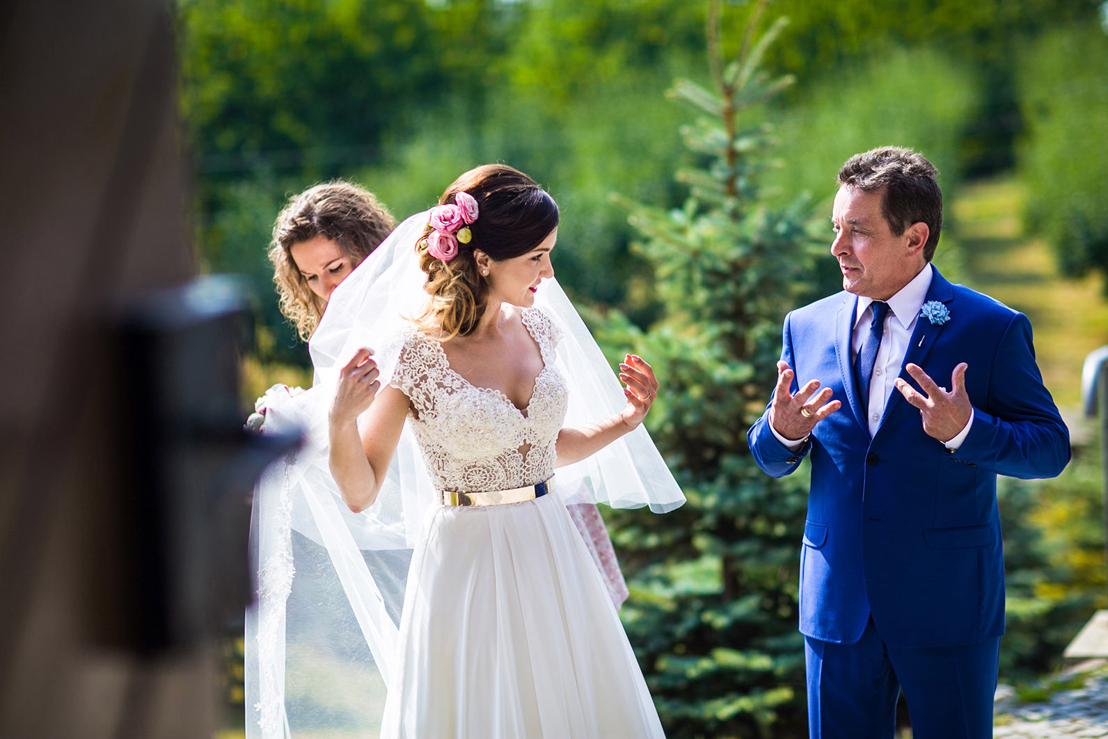 Panna młoda w koronkowej sukni z welonem, poprawianym przez druhnę, oraz mężczyzna w niebieskim garniturze na tle zieleni, Wrocław. Ujęcie z przygotowań do ślubu.