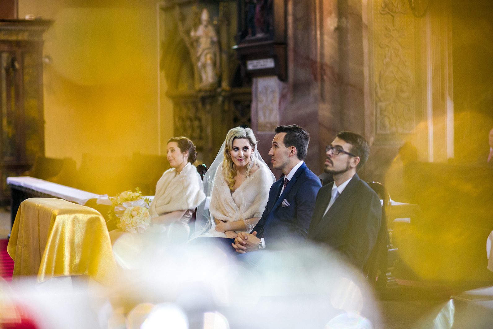 Para młoda w kościele podczas ceremonii ślubnej, otoczona gośćmi. Panna młoda w welonie i futerku, pan młody w granatowym garniturze. Dekoracje kwiatowe i sakralne tło.
