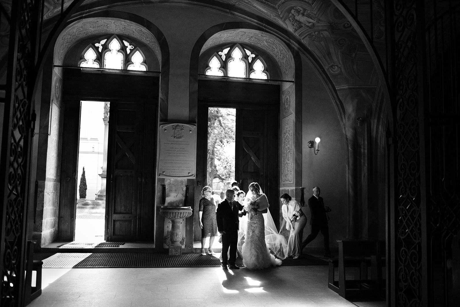 Czarno-białe zdjęcie nowożeńców wychodzących z kościoła, otoczonych gośćmi. Kontrastowe światło podkreśla architektoniczne detale wnętrza.