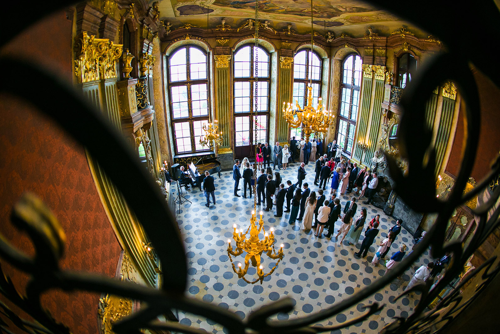 Eleganckie wnętrze z grupą ludzi podczas uroczystości, widok z góry przez ozdobną balustradę. W tle okna i żyrandole, pianista gra na pianinie.