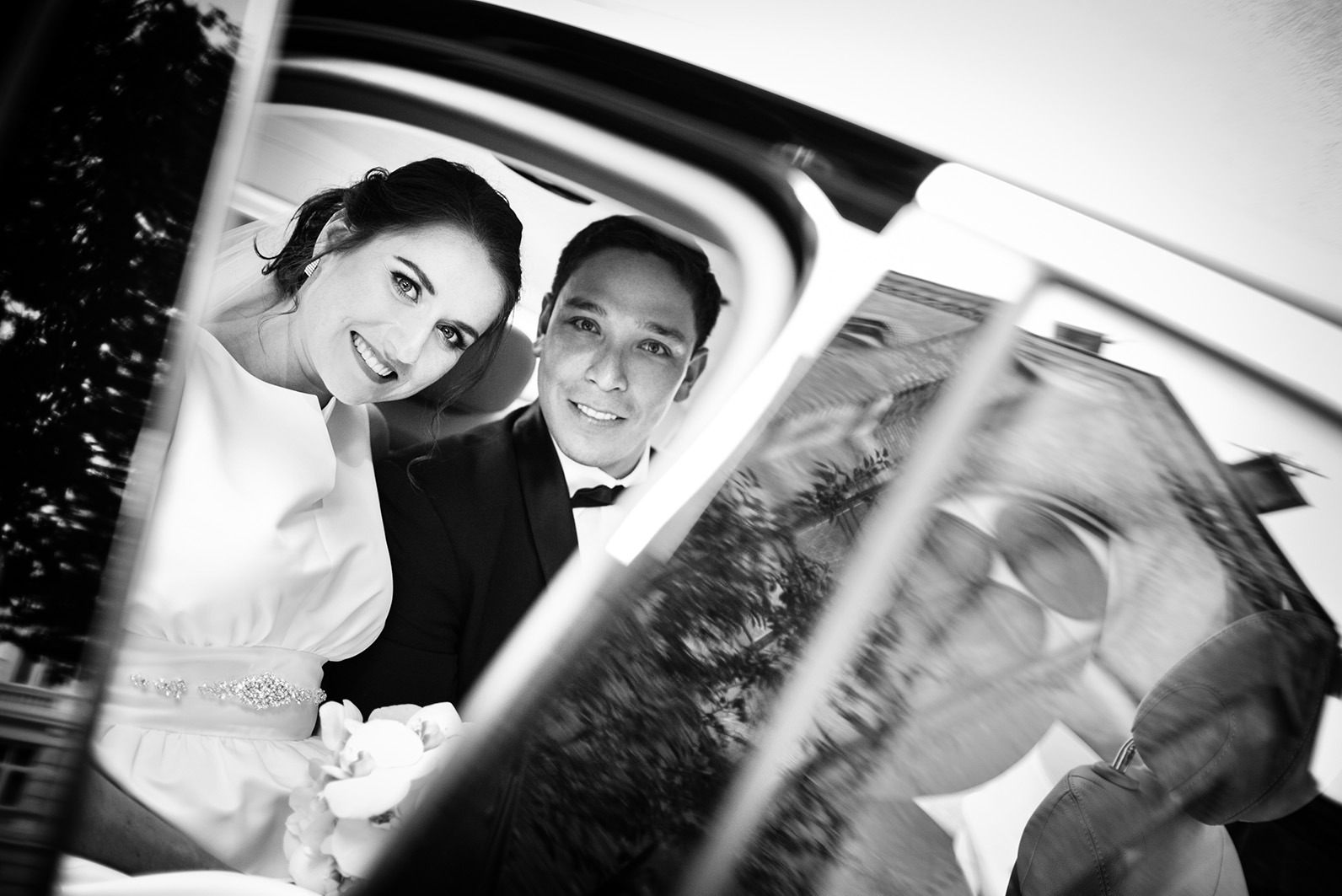 Czarno-białe zdjęcie uśmiechniętej pary młodej w samochodzie, z odbiciem budynku w szybie. Panna młoda w sukni z bukietem, pan młody w smokingu.