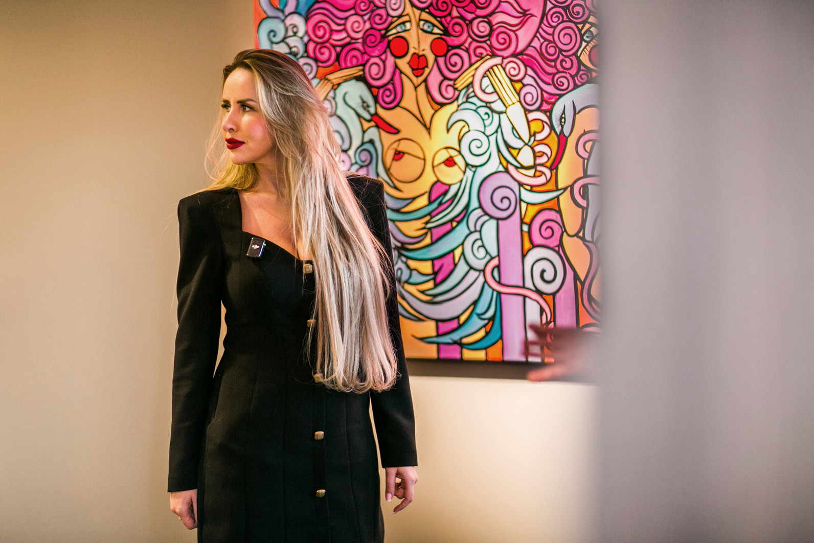 Elegancka blondynka w czarnej sukience pozuje na tle abstrakcyjnego obrazu w żywych kolorach. Profesjonalna sesja w studio, wyrazisty makijaż, spojrzenie w bok.