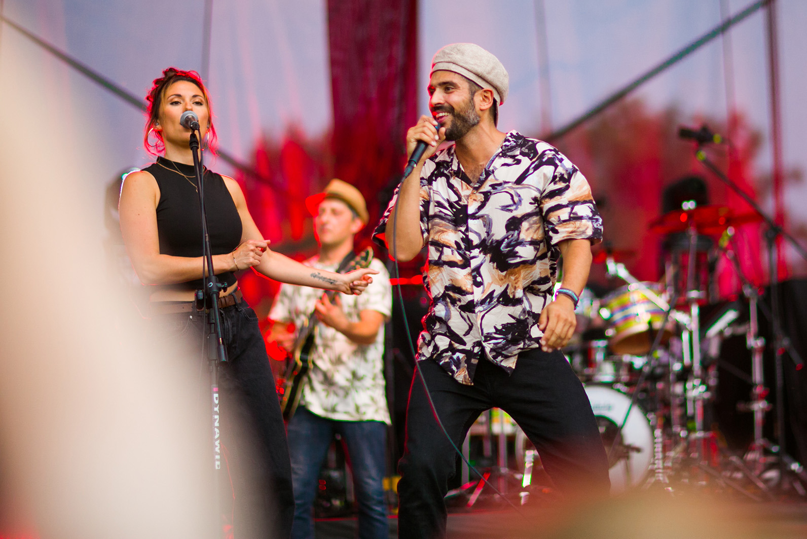 Zespół muzyczny na scenie podczas koncertu: wokalistka z mikrofonem, gitarzysta i perkusista w tle, dynamiczne oświetlenie.