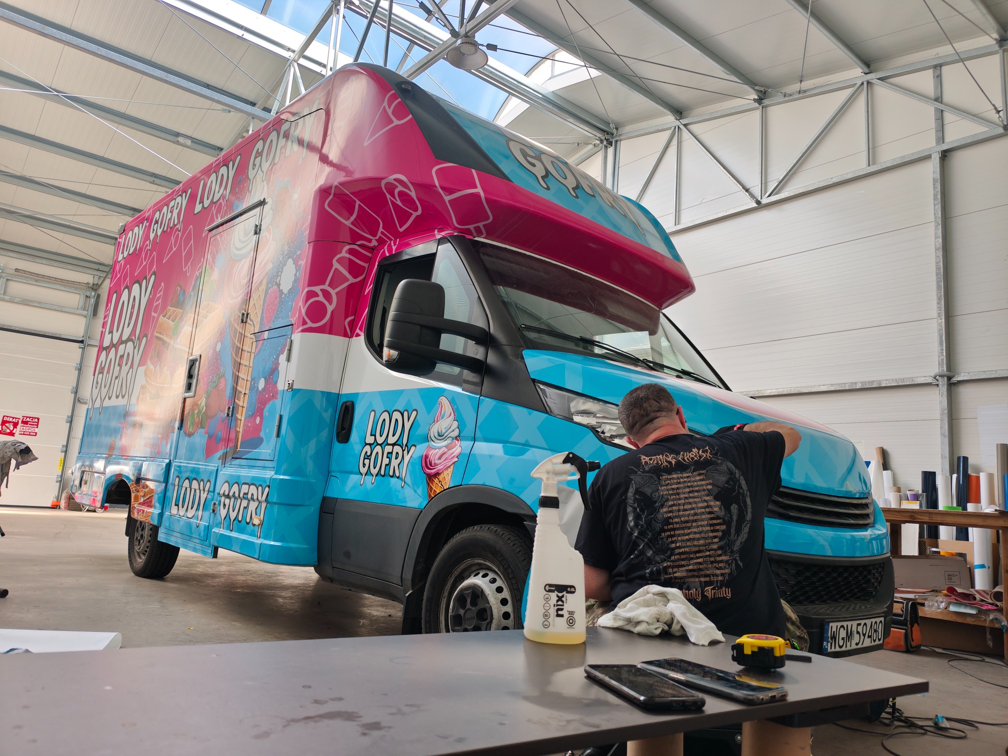Oklejanie food trucka we Wrocławiu. Pracownik aplikuje folię z motywem lodów i gofrów na samochód dostawczy w hali. Widoczne narzędzia i butelka ze spryskiwaczem.