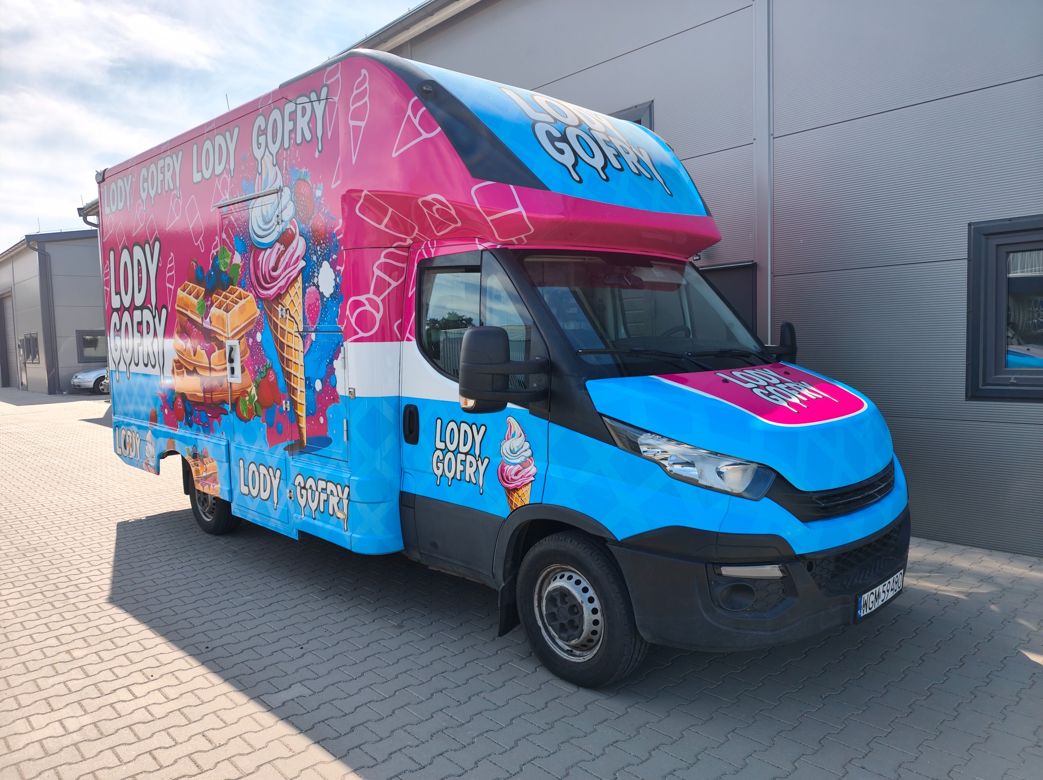 Kolorowy furgon z grafiką lodów i gofrów, zaparkowany na zewnątrz budynku. Jasne, pastelowe barwy i wyraźne napisy przyciągają wzrok. Widoczny numer rejestracyjny.