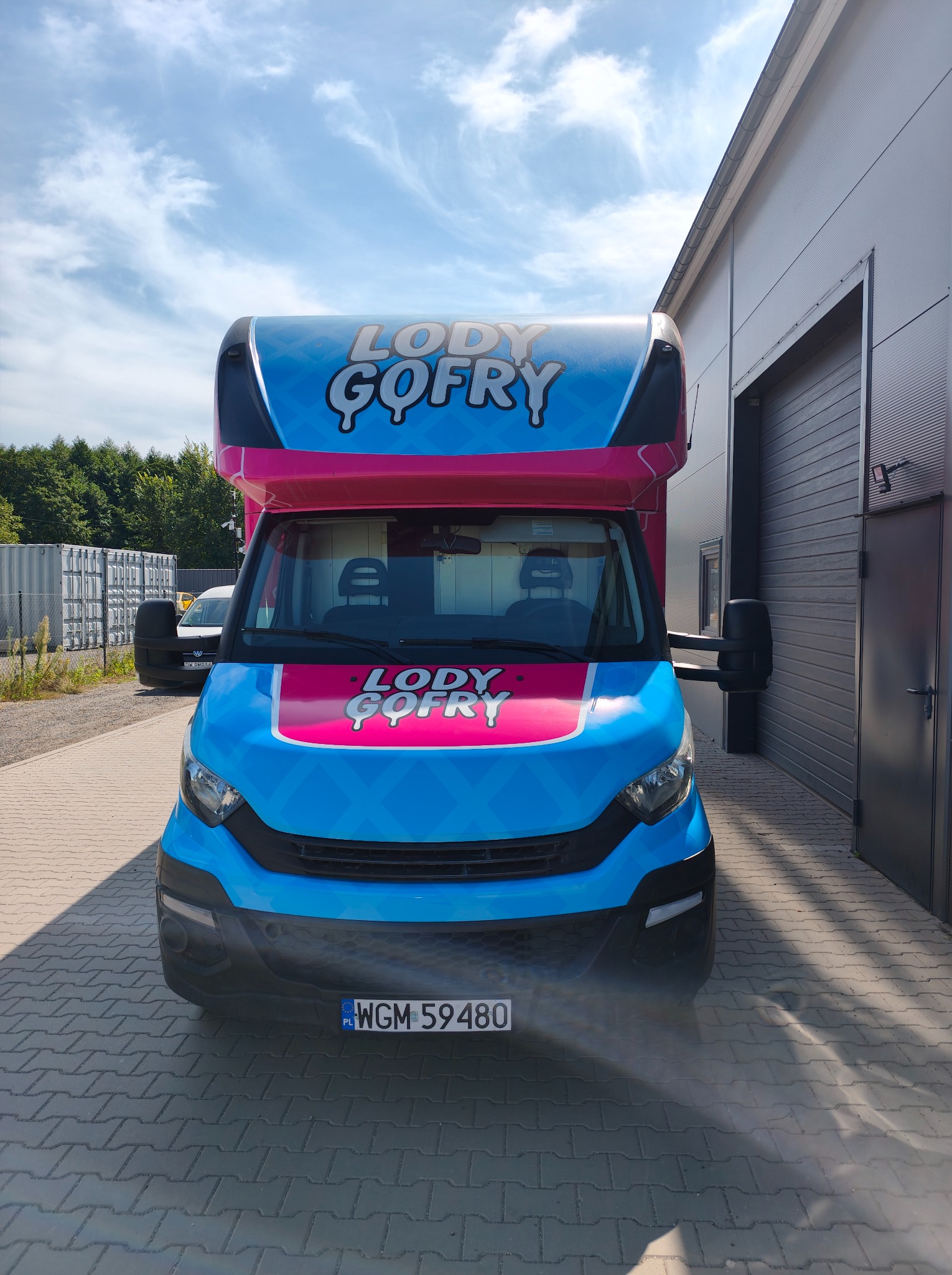 Niebieski food truck z różowymi akcentami i napisem 'Lody Gofry' na tle szarego budynku i błękitnego nieba. Widok z przodu, tablica rejestracyjna WGM 59480.