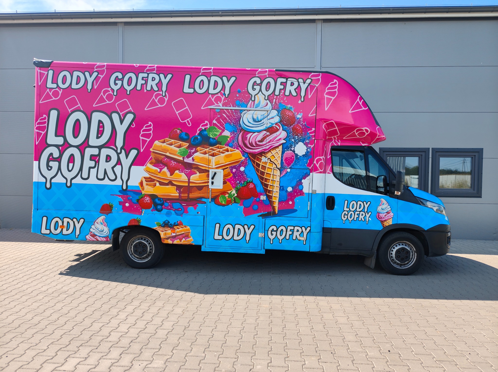Oklejony food truck z grafiką lodów i gofrów w różowo-niebieskiej kolorystyce, zaparkowany na kostce brukowej przed szarą halą. Wyraźne detale grafiki.