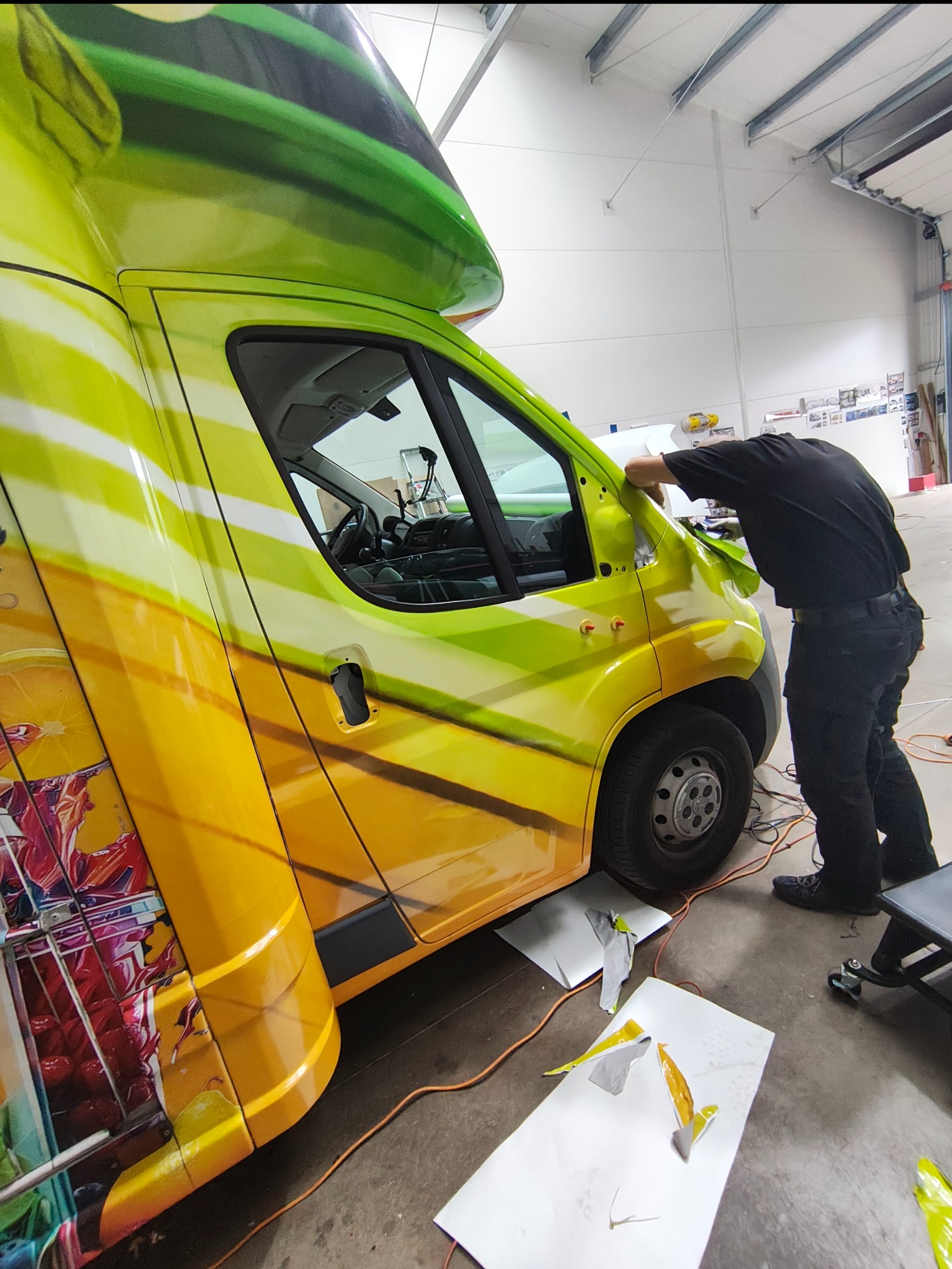 Żółto-zielony van z grafiką owoców, oklejany przez pracownika w czarnym ubraniu. Widoczne narzędzia i resztki folii, co sugeruje etap pracy. Jasne oświetlenie warsztatowe.