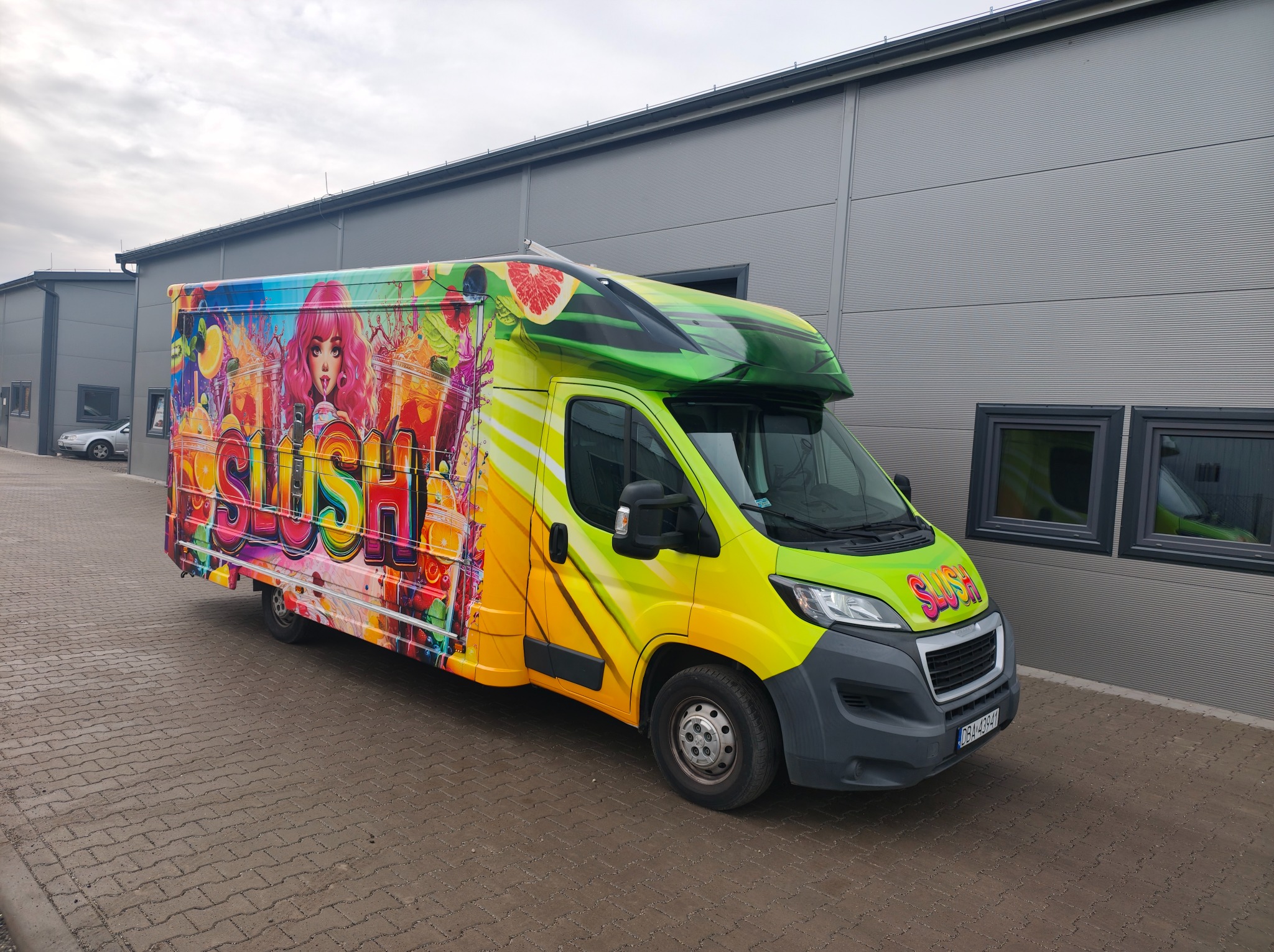 Żółto-zielony van z kolorową grafiką 'SLUSH' i wizerunkiem kobiety, zaparkowany przed szarą halą. Grafika obejmuje całą boczną powierzchnię pojazdu.