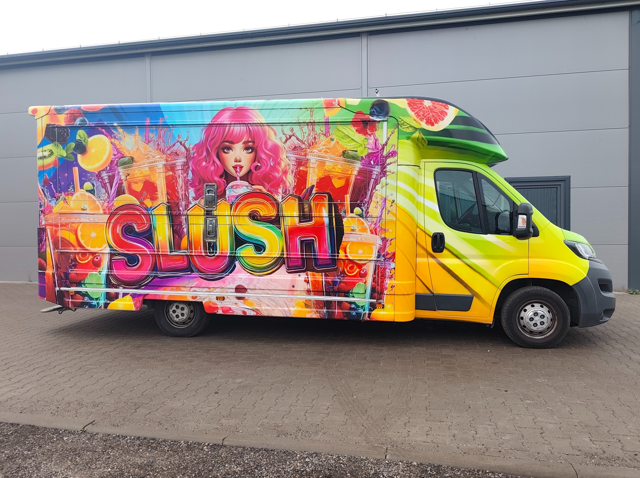Żółty van z grafiką 'SLUSH' i ilustracją dziewczyny z różowymi włosami, otoczonej owocami i napojami, zaparkowany na tle szarego budynku.