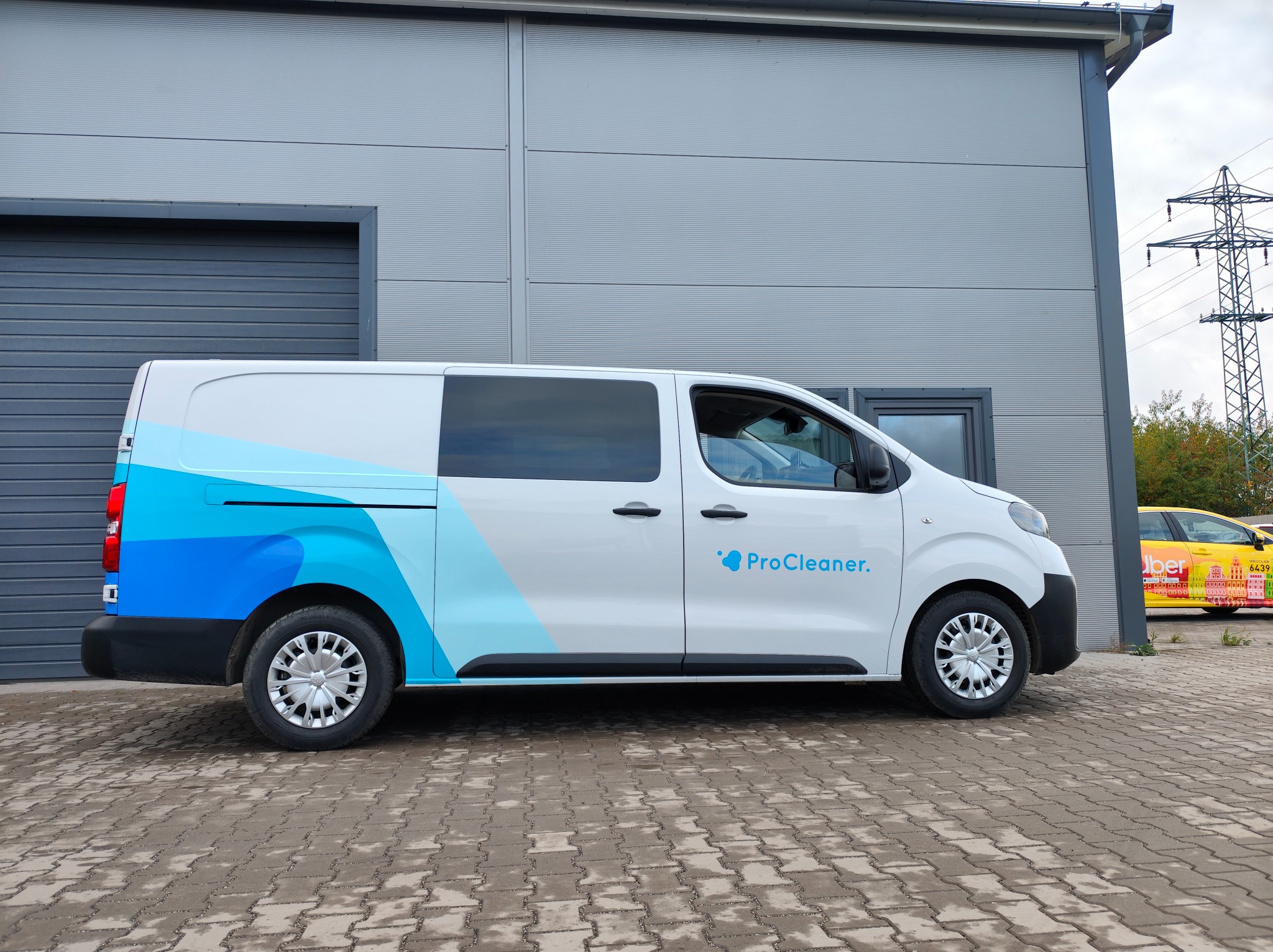Biały van z niebieską grafiką i logo 'ProCleaner' na tle szarego budynku przemysłowego. Widoczny fragment żółtego auta z napisem 'Uber' w tle.