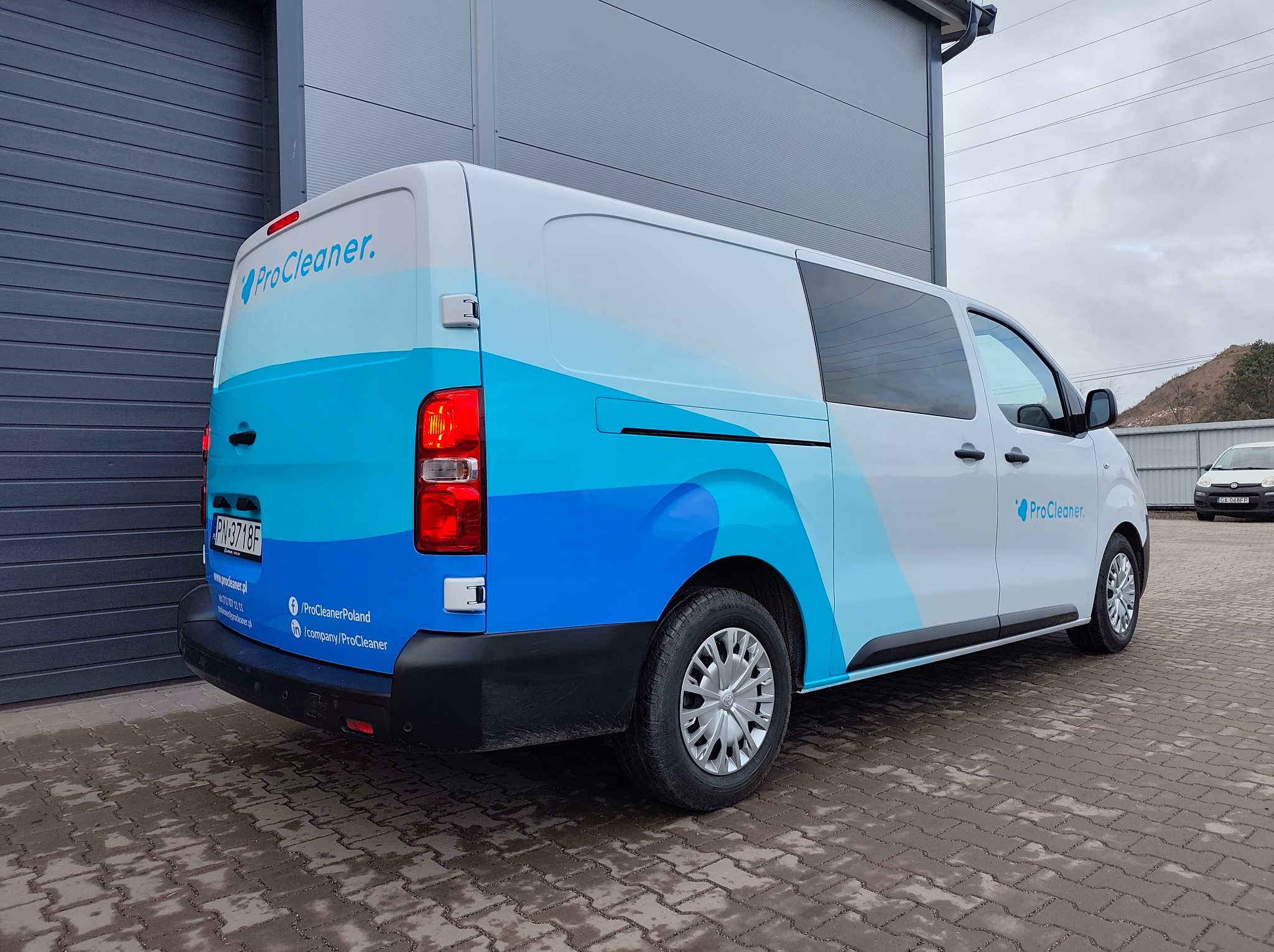 Biały van z niebieską grafiką fal i logo ProCleaner, zaparkowany na kostce brukowej, z widocznym numerem rejestracyjnym i danymi kontaktowymi firmy.