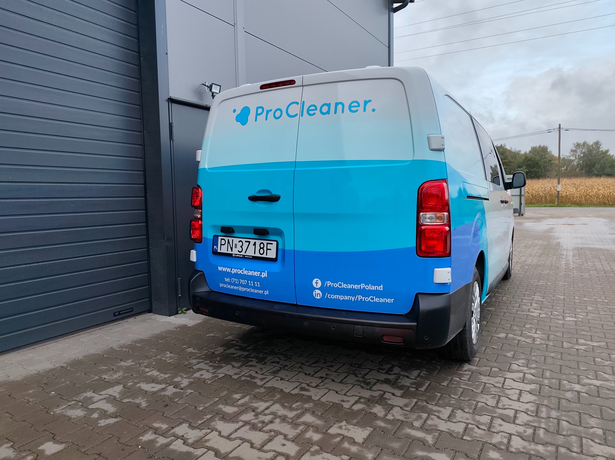 Biały van z błękitnym pasem i logo ProCleaner zaparkowany na mokrym bruku, widok z tyłu. Na karoserii dane kontaktowe firmy. W tle budynek przemysłowy i pole kukurydzy.