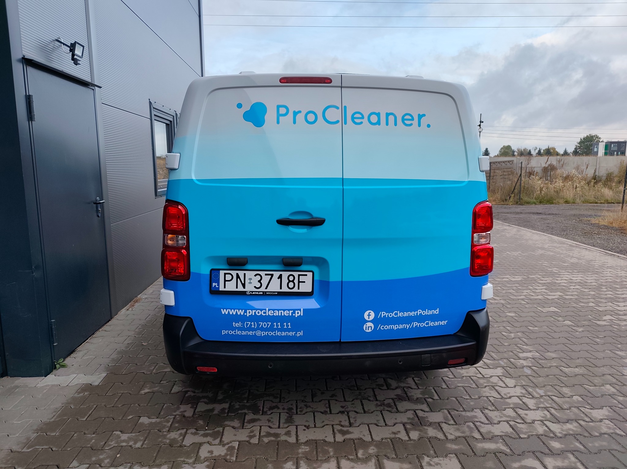 Oklejony na niebiesko-biało van firmy ProCleaner z widocznym numerem rejestracyjnym i danymi kontaktowymi, zaparkowany na kostce brukowej przed szarą halą.