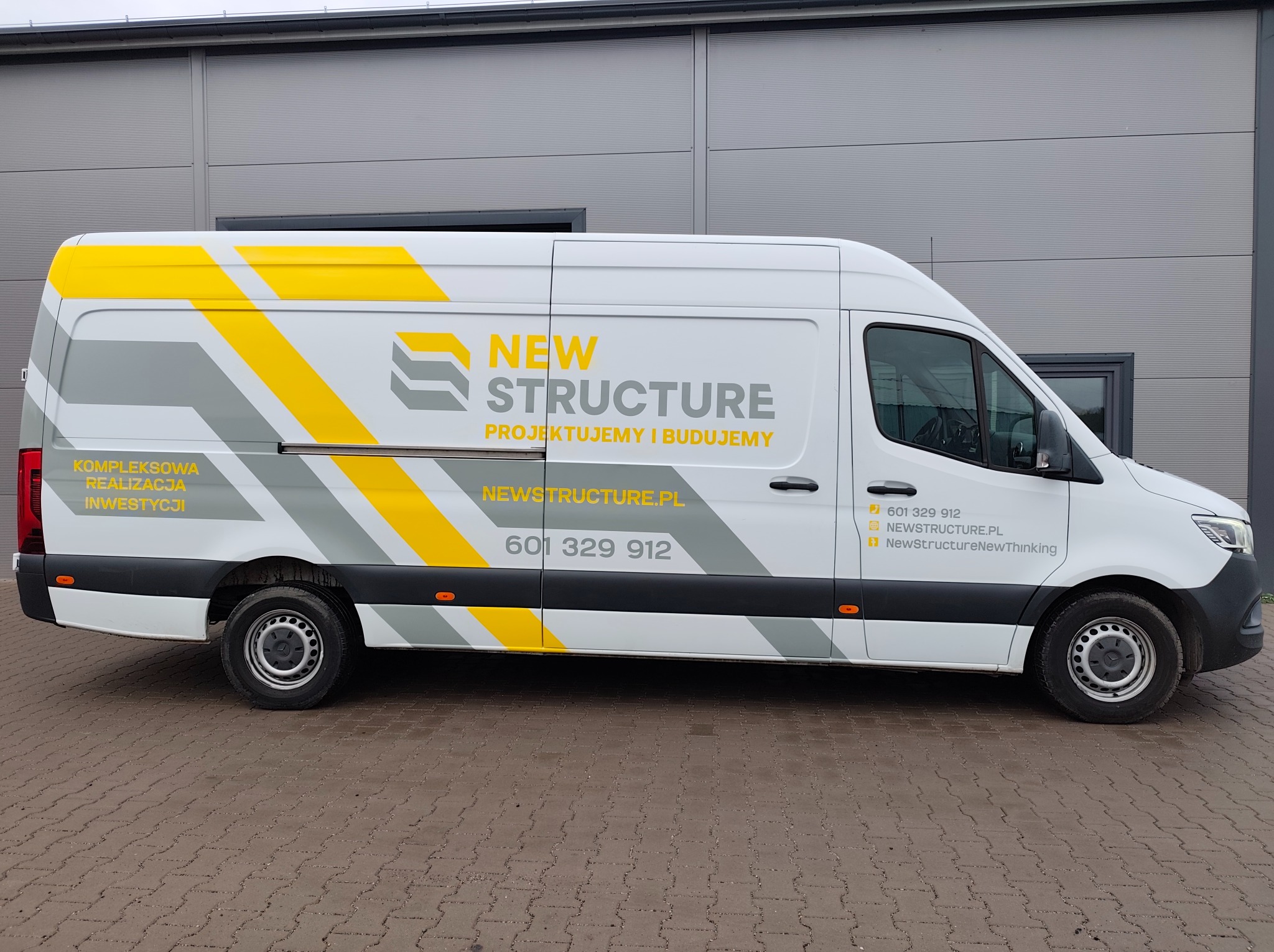 Biały van z szaro-żółtą grafiką i logo firmy New Structure na boku, zaparkowany na kostce brukowej przed szarym budynkiem. Prezentacja oklejonego pojazdu.