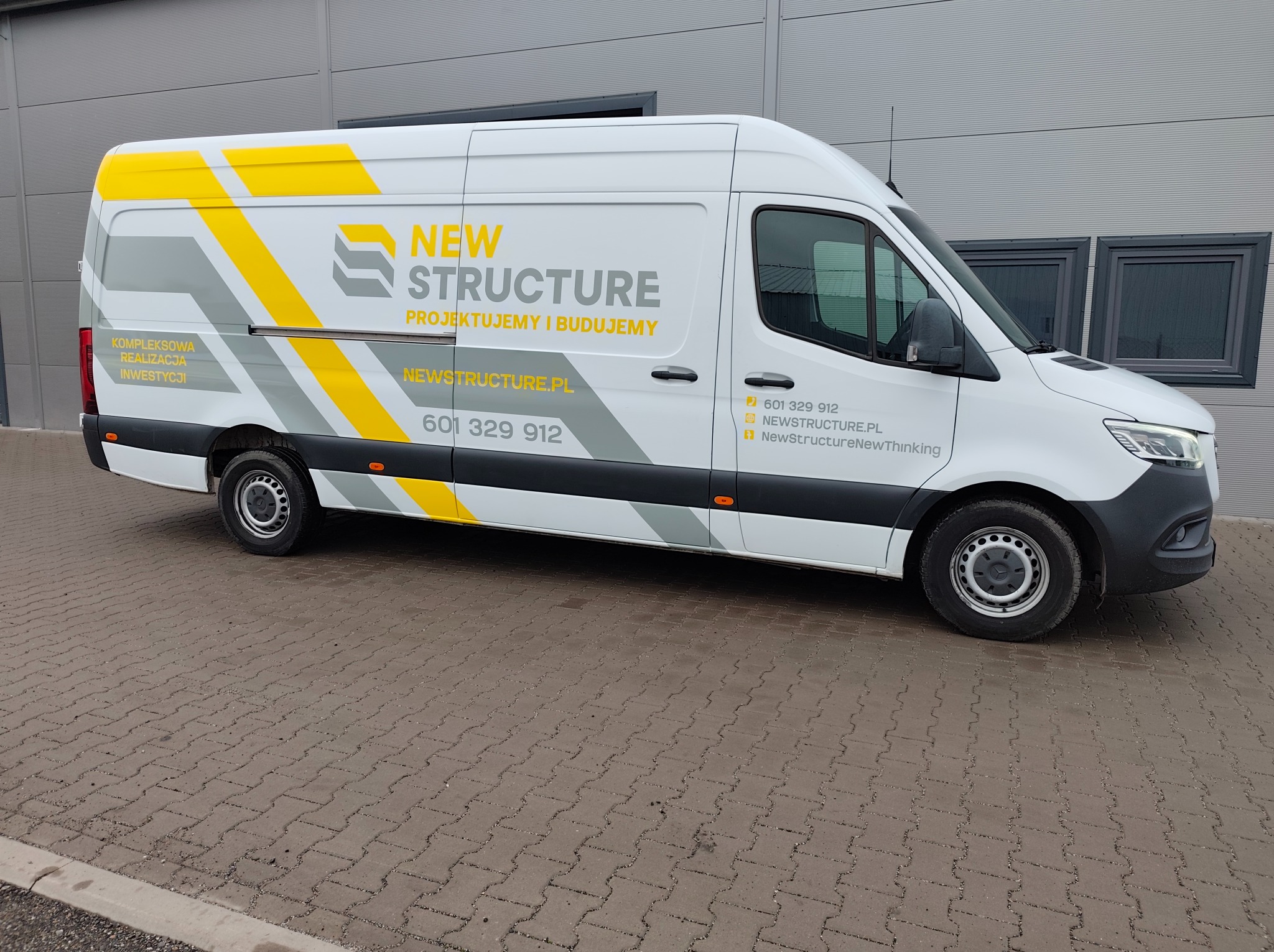 Biały van z żółto-szarymi elementami graficznymi i logo firmy budowlanej 'New Structure' na boku, zaparkowany na brukowanej powierzchni przed szarym budynkiem.
