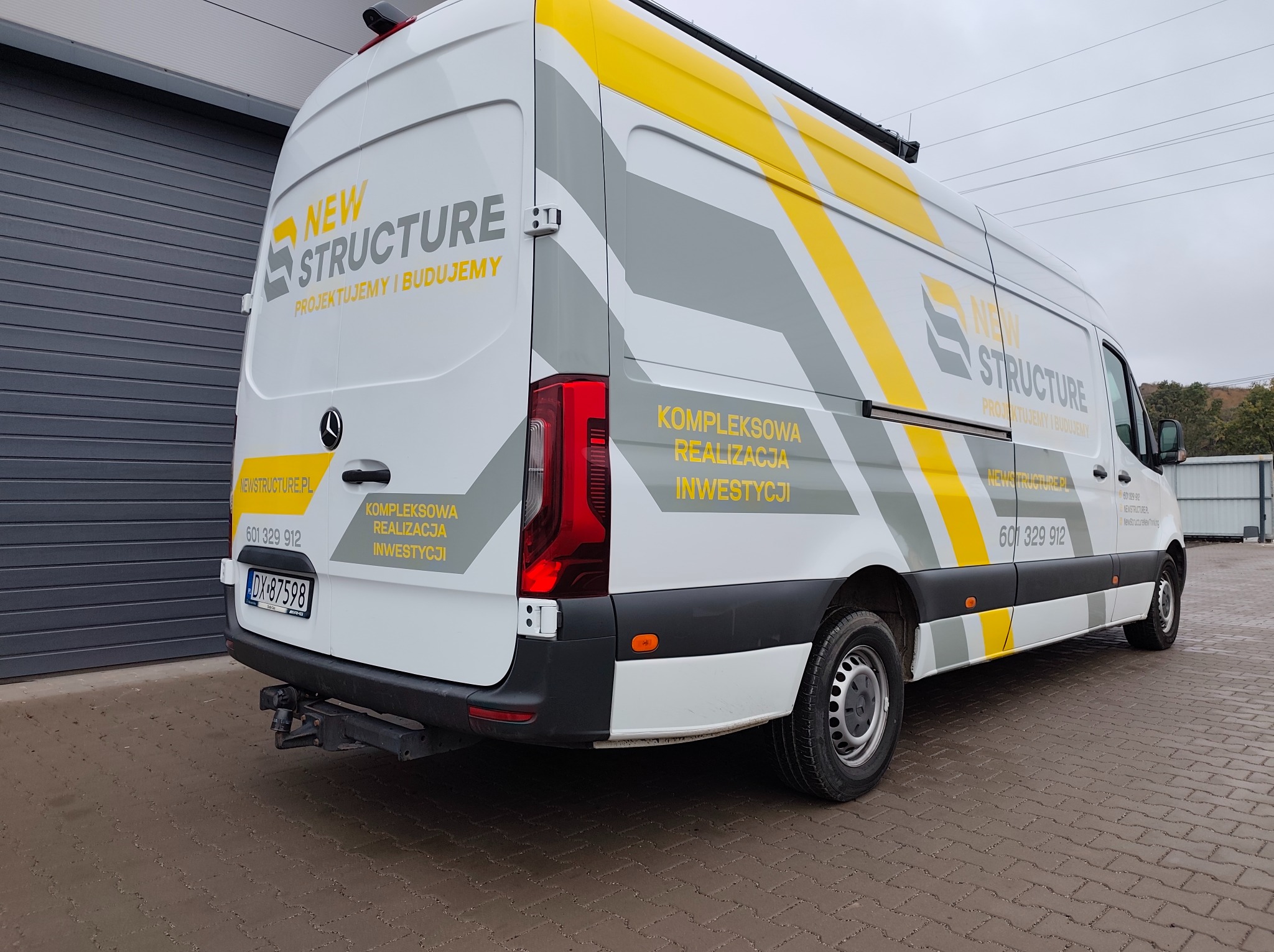 Biały van z szaro-żółtymi pasami i logo firmy budowlanej 'New Structure' na boku, zaparkowany na brukowanej nawierzchni, z widocznym hakiem holowniczym.