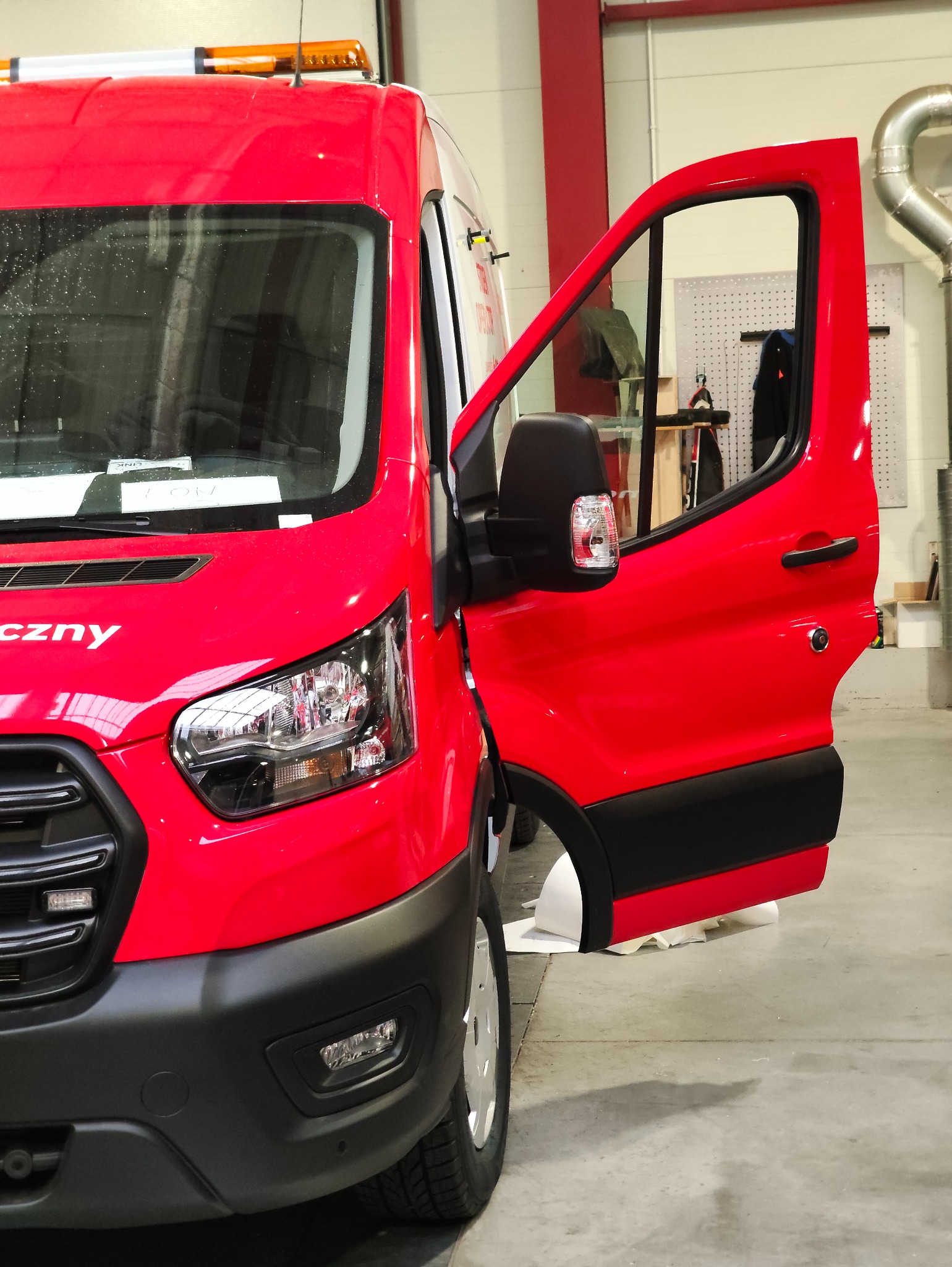 Czerwony bus z otwartymi drzwiami w warsztacie. Widoczny fragment okleiny i detale karoserii. Oświetlenie warsztatowe.