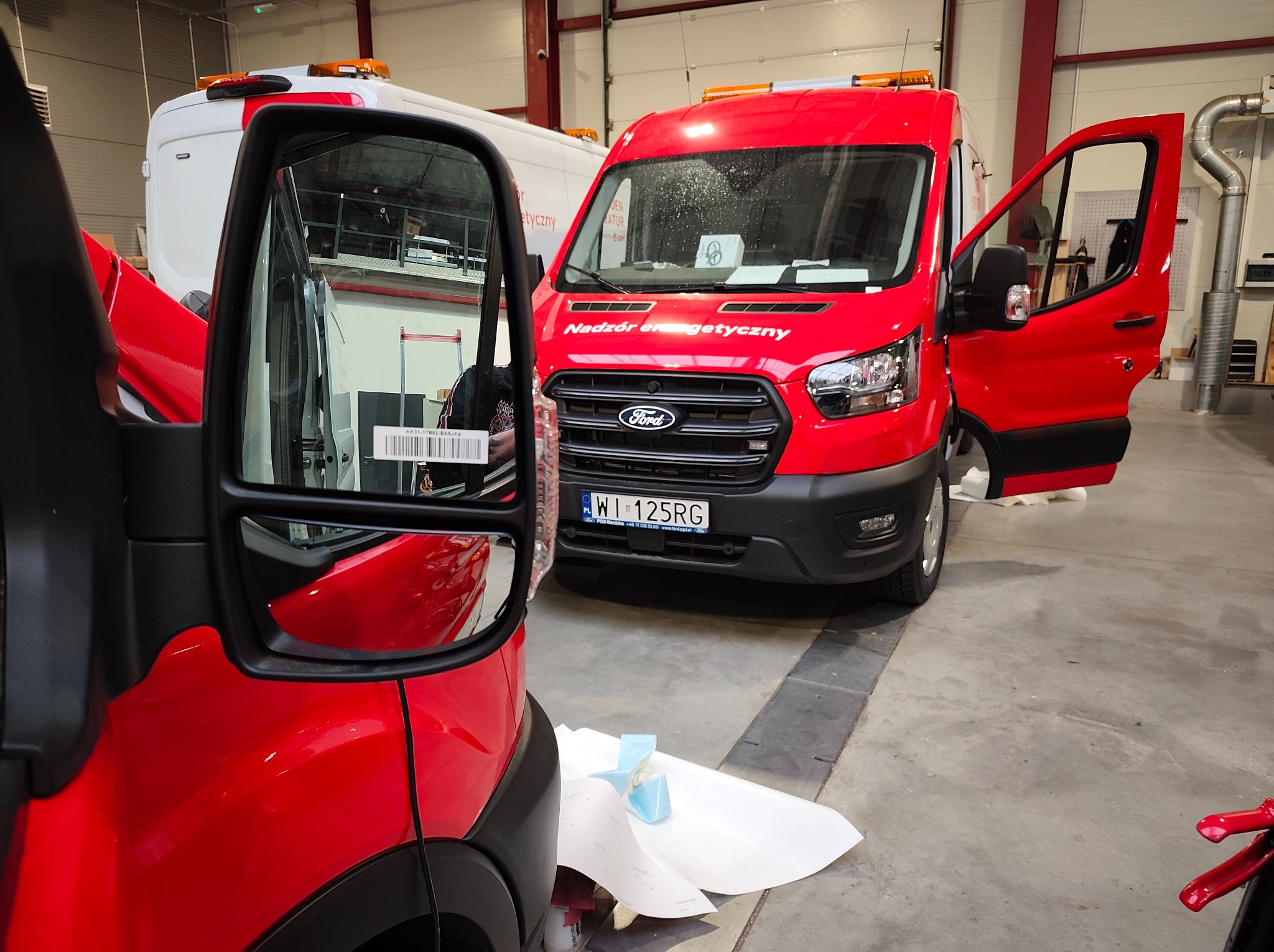 Czerwony bus z otwartymi drzwiami i napisem 'Nadzór energetyczny', widoczny w warsztacie. Fragment okleiny na podłodze sugeruje proces aplikacji.