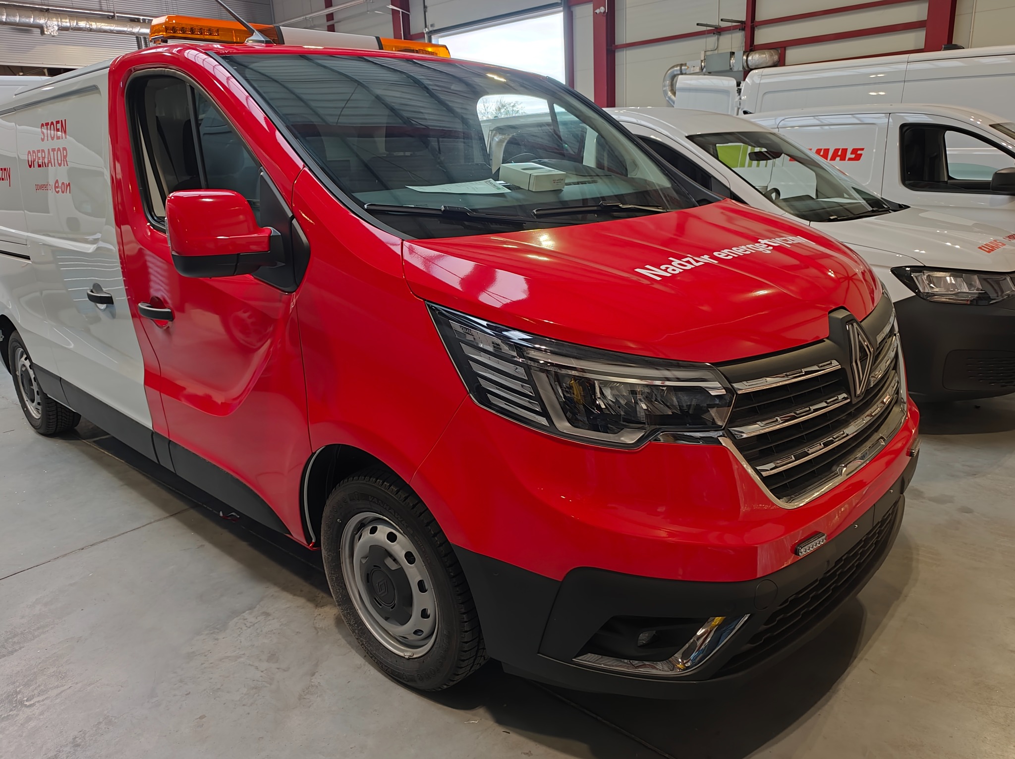 Czerwono-biały van z napisem 'Nadzór energetyczny' i lampą ostrzegawczą na dachu, zaparkowany w hali. Widoczne inne pojazdy w tle.