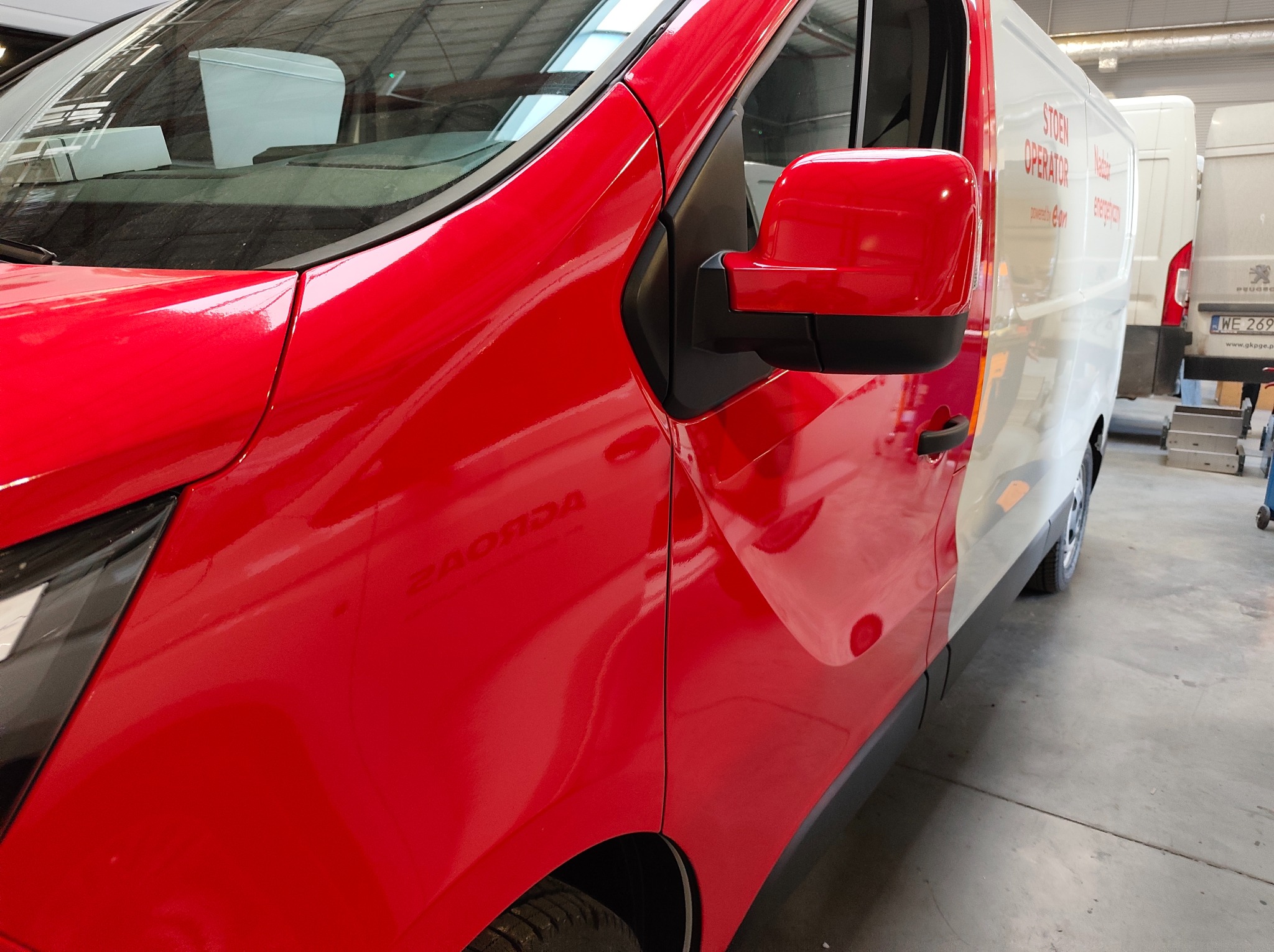 Czerwono-biały van po oklejeniu, widoczny fragment karoserii z lusterkiem. Na białej części nadrukowane logo firmy. Częściowo widoczny inny pojazd w tle.